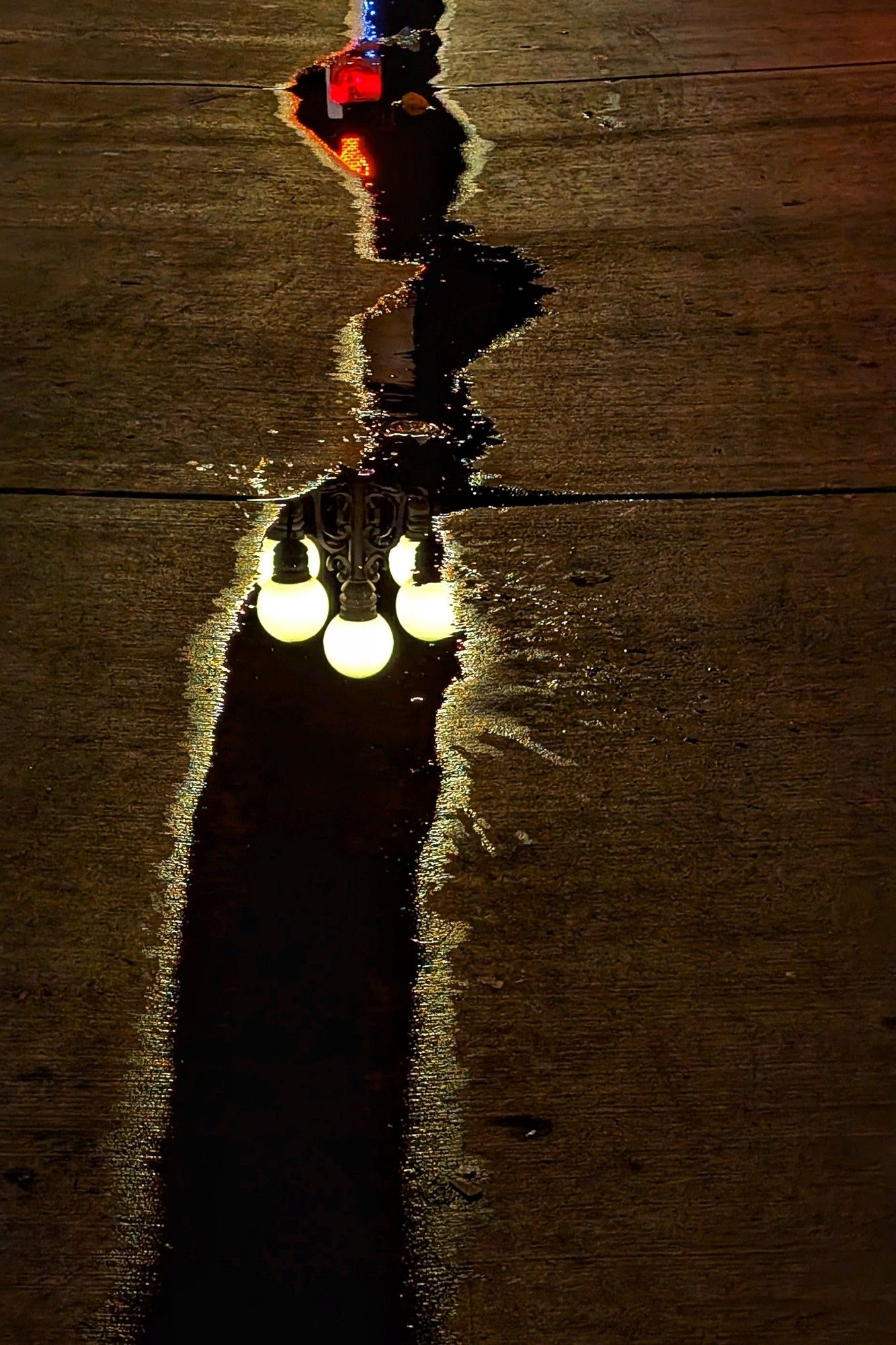 Eine langgezogene Pfütze bei Nacht auf gold schimmerndem Asphalt. In der Pfütze spiegelt sich eine Straßenlaterne, die aus mehreren runden Lampen besteht, sowie eine rote Ampel.