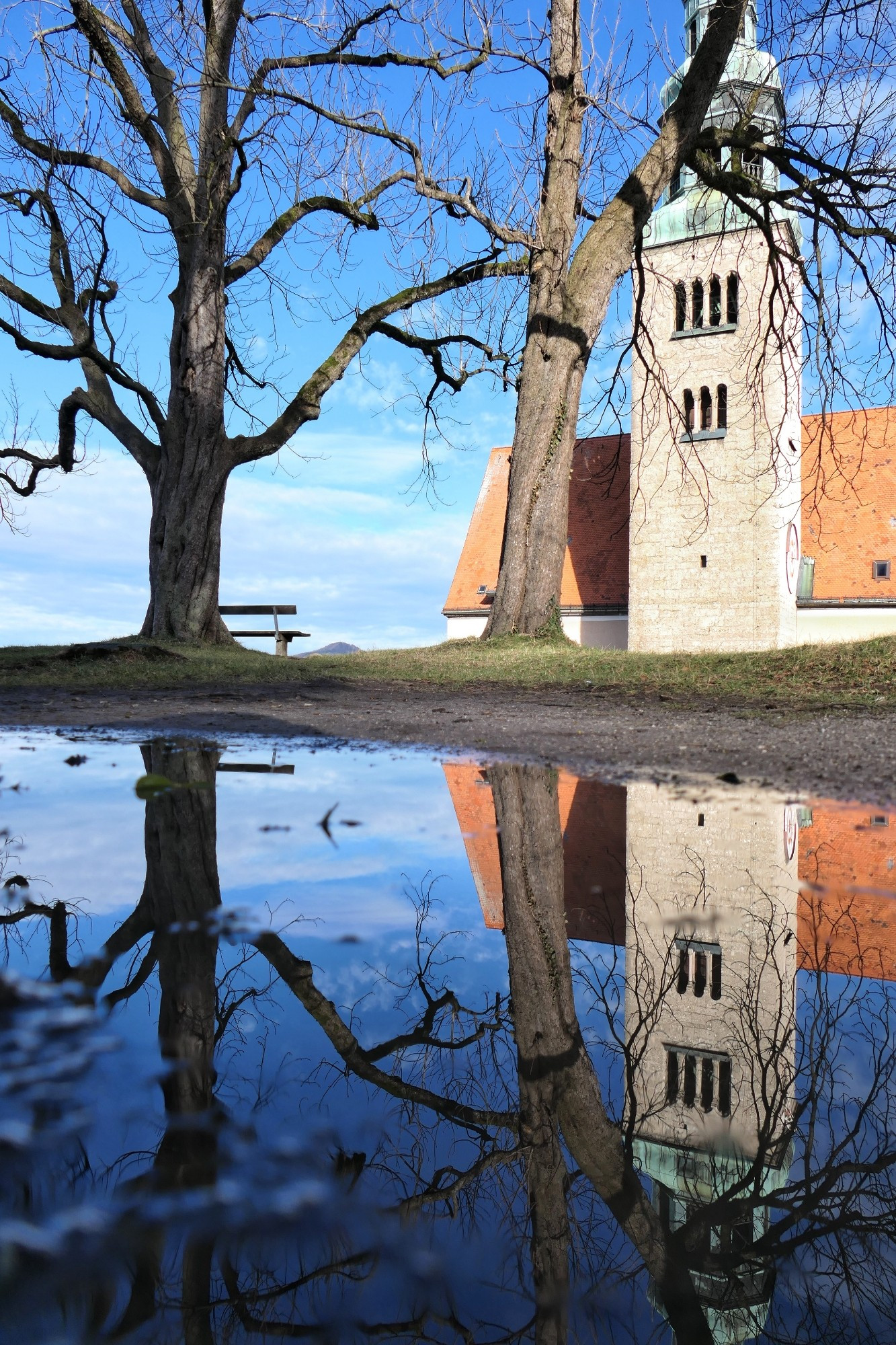 Das Bild zeigt eine Kirche mit einem hohen Turm, zwei Bäume und eine Bank, die sich alle in einer Pfütze spiegeln. Die Kirche hat einen hohen, schlanken Turm und ein rotes Dach. Ein großer kahler Baum links steht auf einem Hügel neben der Kirche; seine Äste sind weit verbreitet. Ein weiterer Baum steht zwischen dem Baum links und dem Kirchturm. Eine hölzerne Bank ist neben dem linken Baum positioniert. Der Himmel ist blau mit wenigen Wolken. Es gibt eine große Pfütze auf dem Boden, in der sich die Kirche, die Bäume und die Bank spiegeln, was eine malerische Ansicht bietet. (Bing-Beschreibung, angepasst)