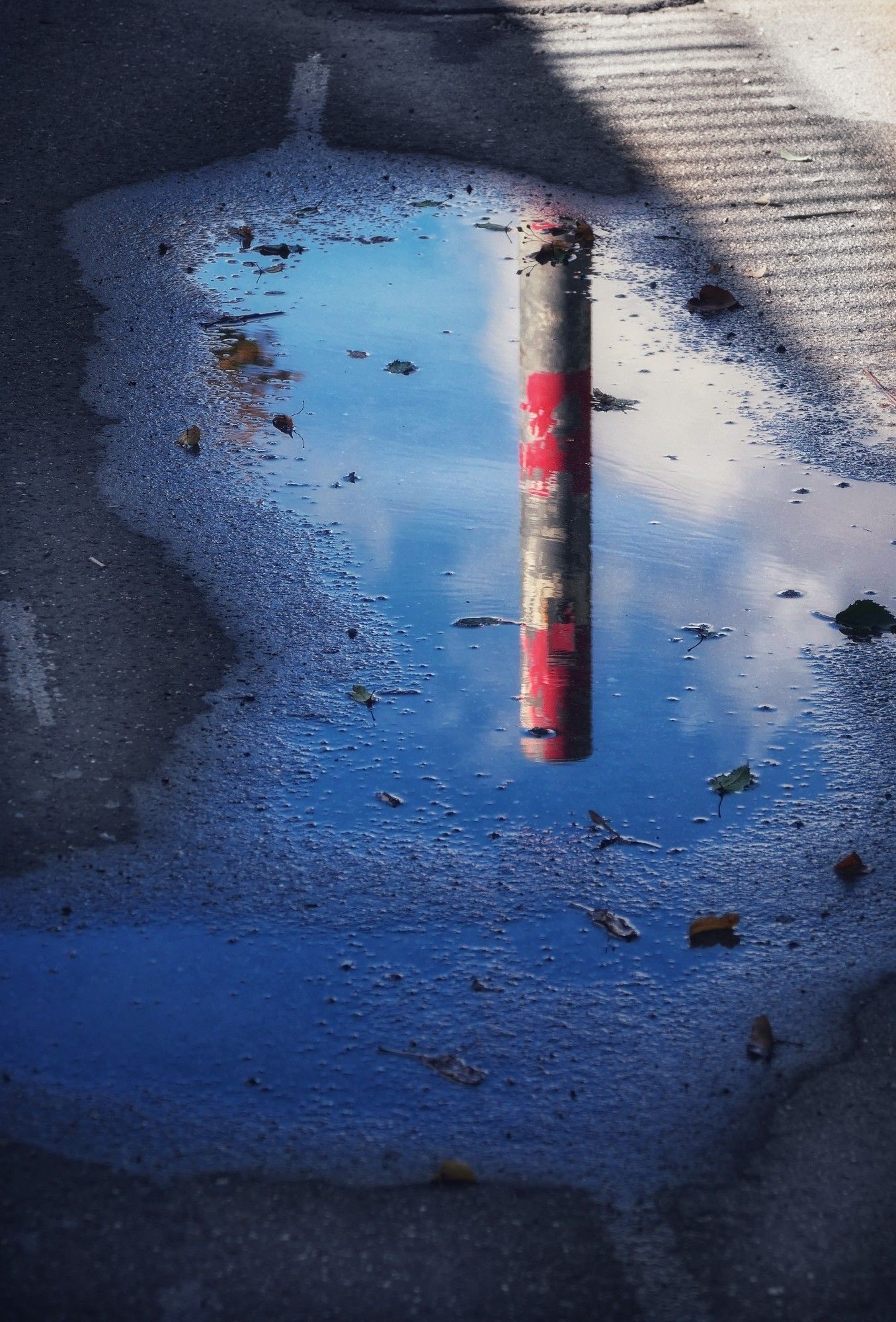 Eine Pfütze, in der sich ein rot-weißer Poller vor blau-weißem Himmel spiegelt.