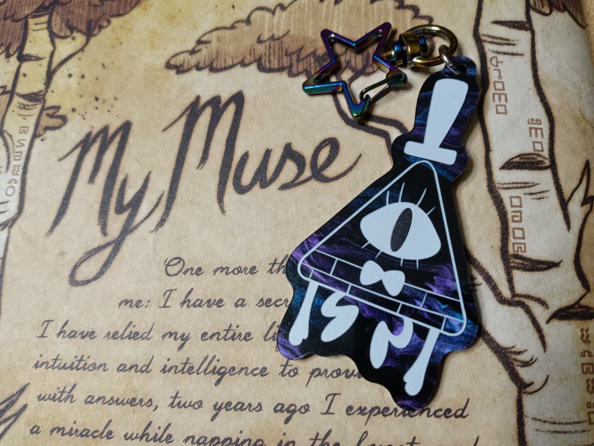 Charm of Bill Cipher from the show Gravity Falls. The art is a white ink outline over black blue and purple swirled acrylic that looks like a galaxy. The background features a journal page that says "My Muse".