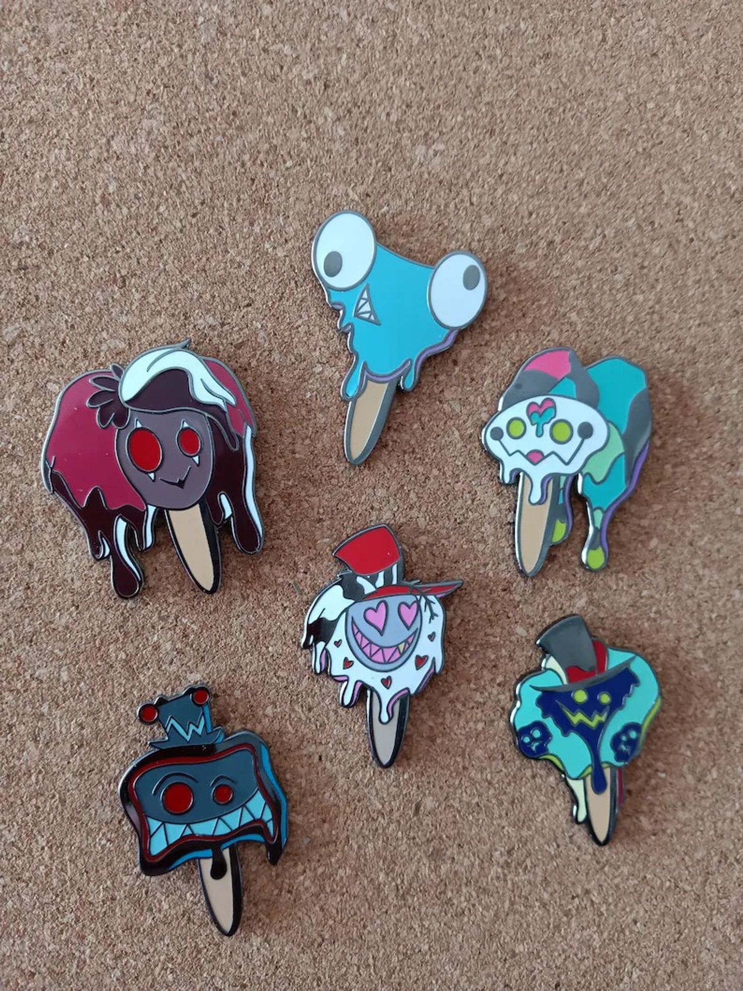 Enamel pins on a corkboard featuring characters from Hazbin Hotel and Helluva Boss