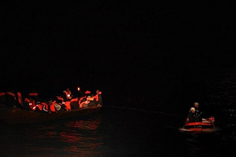 Dunkle Nacht auf See. Ein kleines überfülltes Boot. Dicht gedrängt sitzen die Menschen darauf. Ihre Rettungswesten leuchten in der Dunkelheit. Daneben ein kleines RHIB mit 2 Personen darauf.