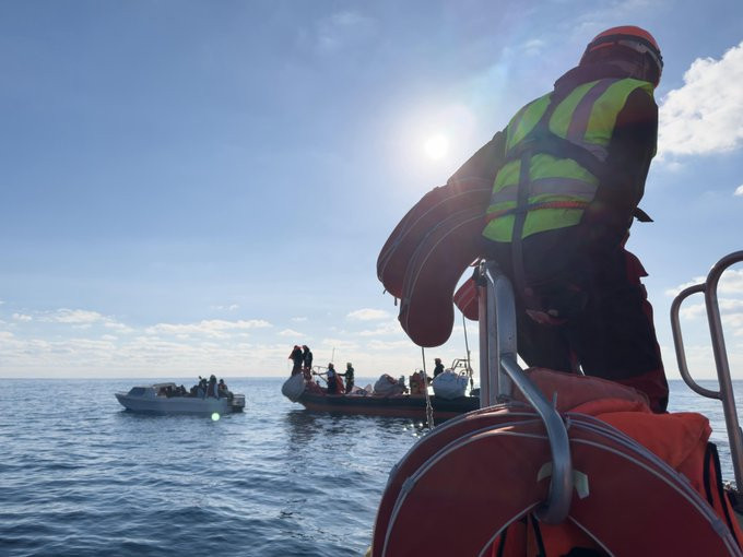 Eine Person auf einem RHIB mit einem Rettungskragen im Arm beobachtet, wie sich ein weiteres RHIB vorsichtig einem kleinen weißen Boot nähert, auf dem Menschen dicht zusammen stehen.