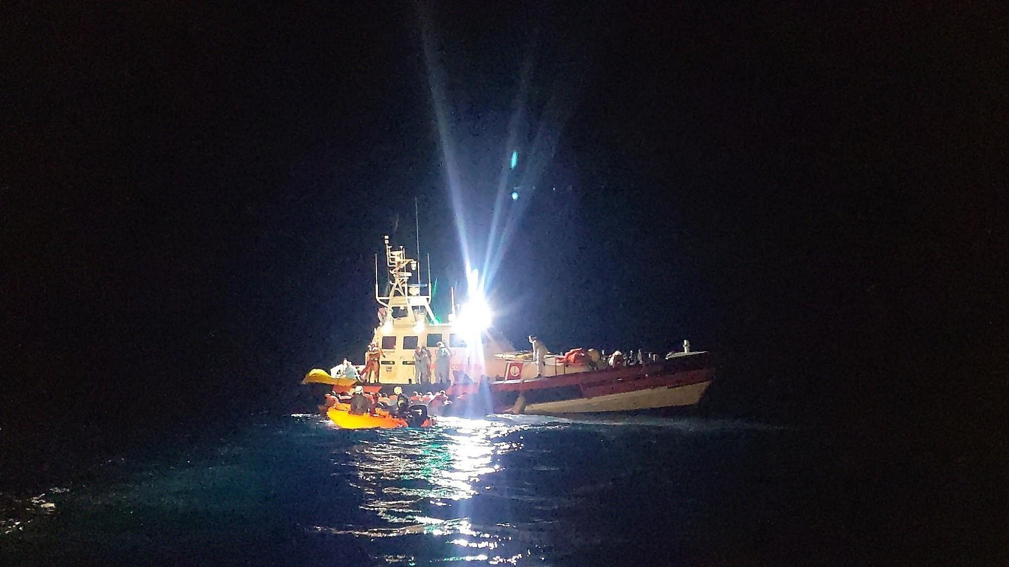 Dunkle Nacht auf See. Eine CP der Guardia Costiera mit hell leuchtenden Scheinwerfern liegt längsseits eines kleinen Bootes voller Menschen. Ein kleines RHIB sichert die Situation auf der anderen Seite des Bootes.