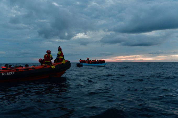 Auf Wasserhöhe aufgenommen: Ein RHIB mit geretteten Menschen an Bord. Im Bug sitzen drei Seenotretter*innen. Dahinter ein immer noch überfülltes GFK-Boot voller Menschen, die mit Rettungswesten versorgt sind.