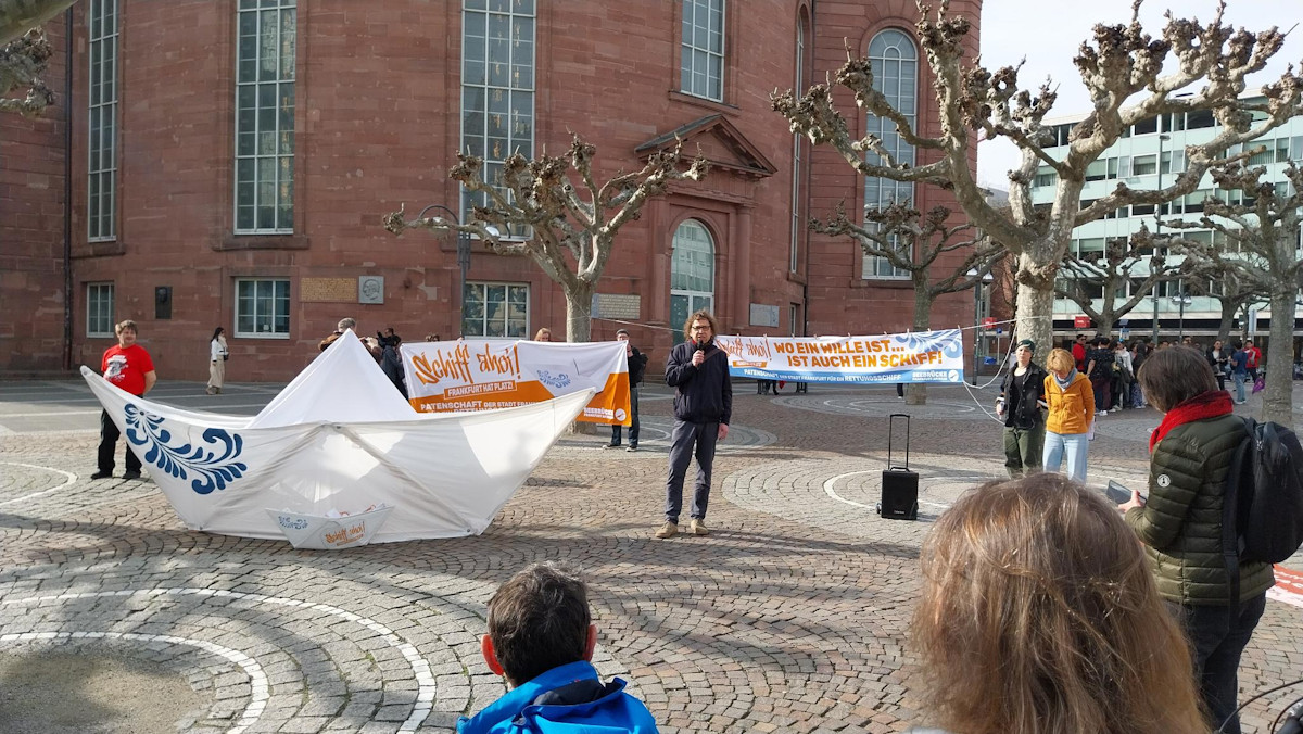 Vor der Paulskirche ist ein überdimensionales Papierfaltboot mit einer blauen Bembelblume aufgebaut. Daneben spricht eine Person in ein Mikrofon. Dahinter zwei Transpis: das orange-weiße Transpi zur Schiff ahoi! Kampagne mit einem weißen Bembelboot. daneben eine Transpi: Wo ein Wille ist, ist auch ein Schiff! Patenschfat der Stadt Frankfurt für ein Rettungsschiff.