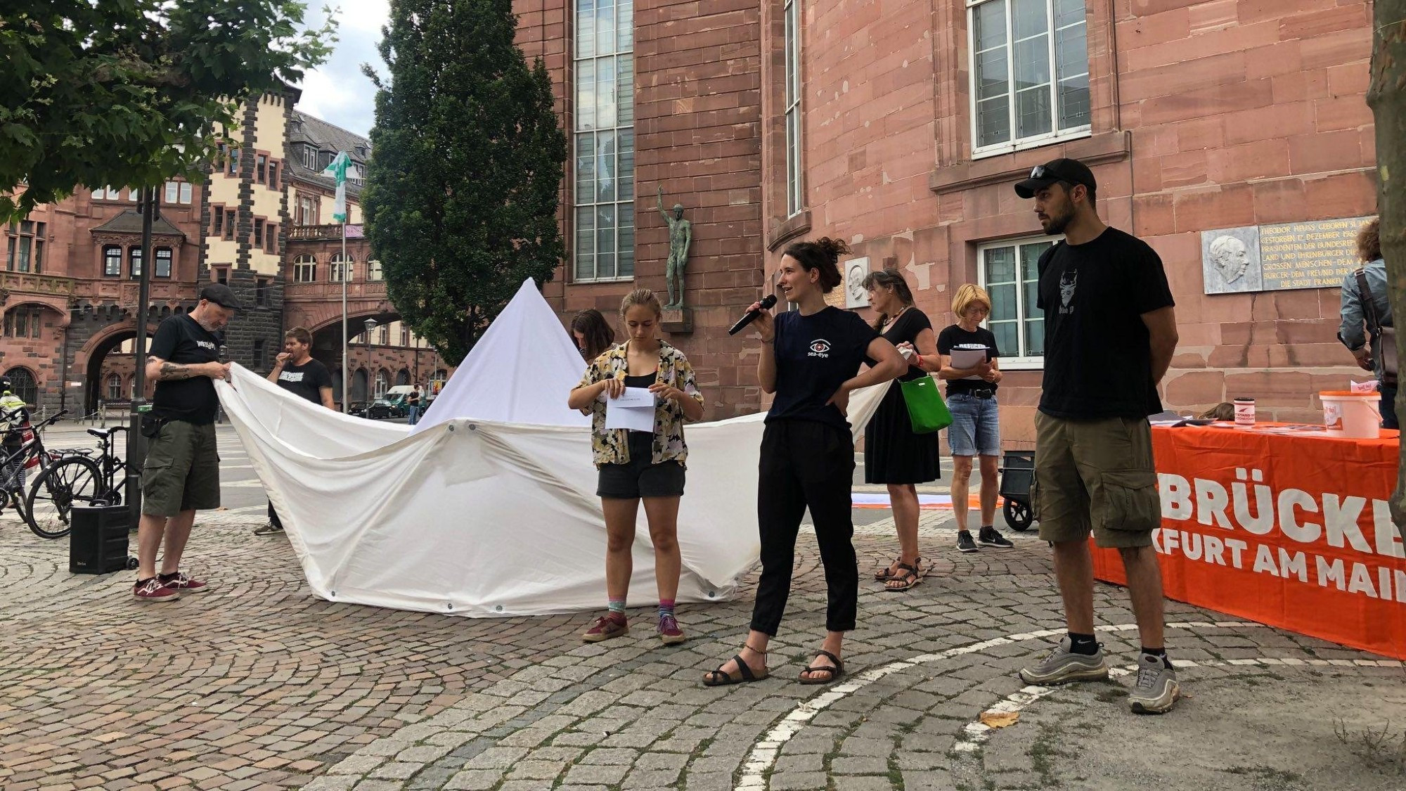 Ein übergroßes Faltschiff vor der Paulskirche. Menschen mit Sea Eye Shirts halten eine Rede.