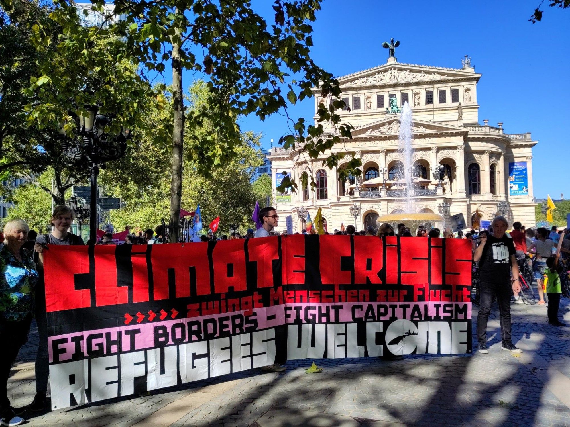 Vor der Alten Oper in strahlendem Sonnenschein Seebrücke mit großem leuchtenden Transpi:
CLIMATE CRISIS
zwingt Menschen zur Flucht

Fight Borders - Fight Capitalism
REFUGEES WELCOME