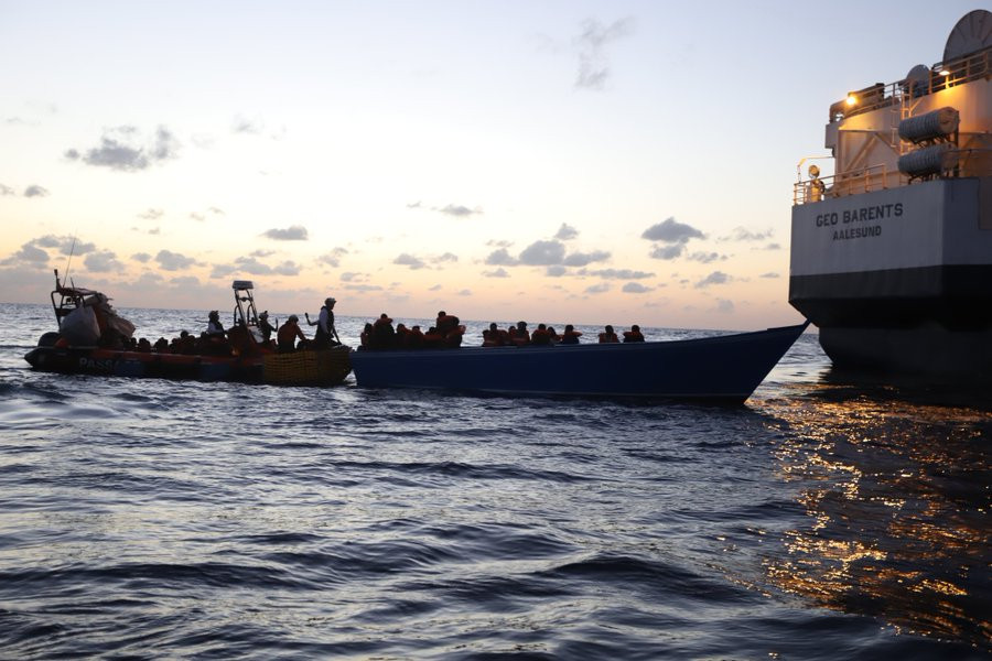 Ein Holzboot, auf dem in der Mitte mehrere Menschen sitzen und am Heck mehrere Personen stehen. Dort hat ein RHIB angedockt, in dem sich schon mehrere gerettete Personen befinden. Rechts ragt das Heck der GEO BARENTS ins Bild.