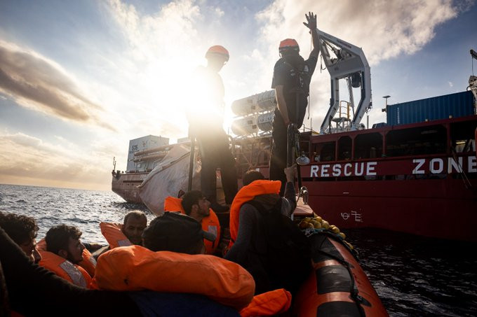 Blick Richtung Bug eines RHIBs. In diesem sitzen Menschen mit Rettungswesten. An der Spitze stehen zwei Seenotretter*innen. Das RHIB nähert sich der OCEAN VIKING. An deren roten Rumpf ist in großen weißen Buchstaben RESCUE ZONE zu lesen.