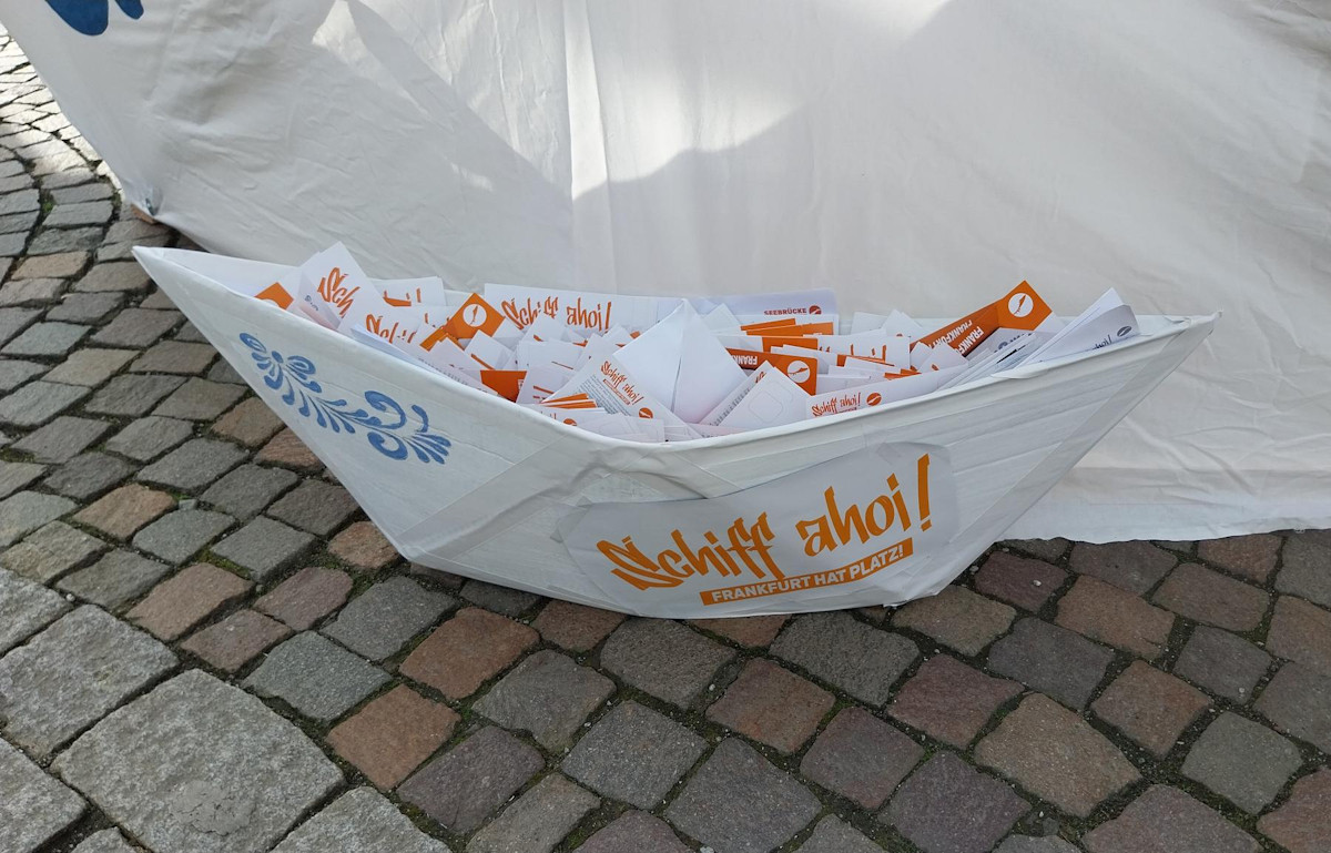 Ein großes Papierfaltboot voller Postkarten. An der Seitenwand eine blaue Bembelblume und in orange: Schiff ahoi! Frankfurt hat Platz