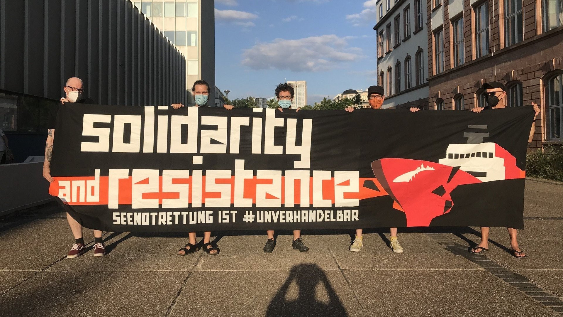 5 Menschen halten ein großes schwarzes Transpi

auf den linken 2/3 steht [Lettern in weiß]:

      solidarity
and resistance [auf einem orangen Streifen]

SEENOTRETTUNG IST #UNVERHANDELBAR

auf dem rechten 1/3 durchbricht ein Schiff mit rotem Rumpf und SB-Ffm-Logo eine schwarze Wand, Brocken fliegen