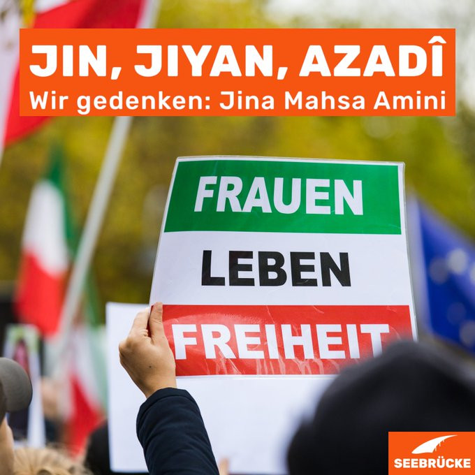 Foto eines Protests, bei dem ein Schild hochgehalten wird, auf dem "Frauen Leben Freiheit" steht. In einem orangen Kasten steht auf dem Foto "Jin, Jiyan, Azadî. Wir gedenken Jina Mahsa Amini"