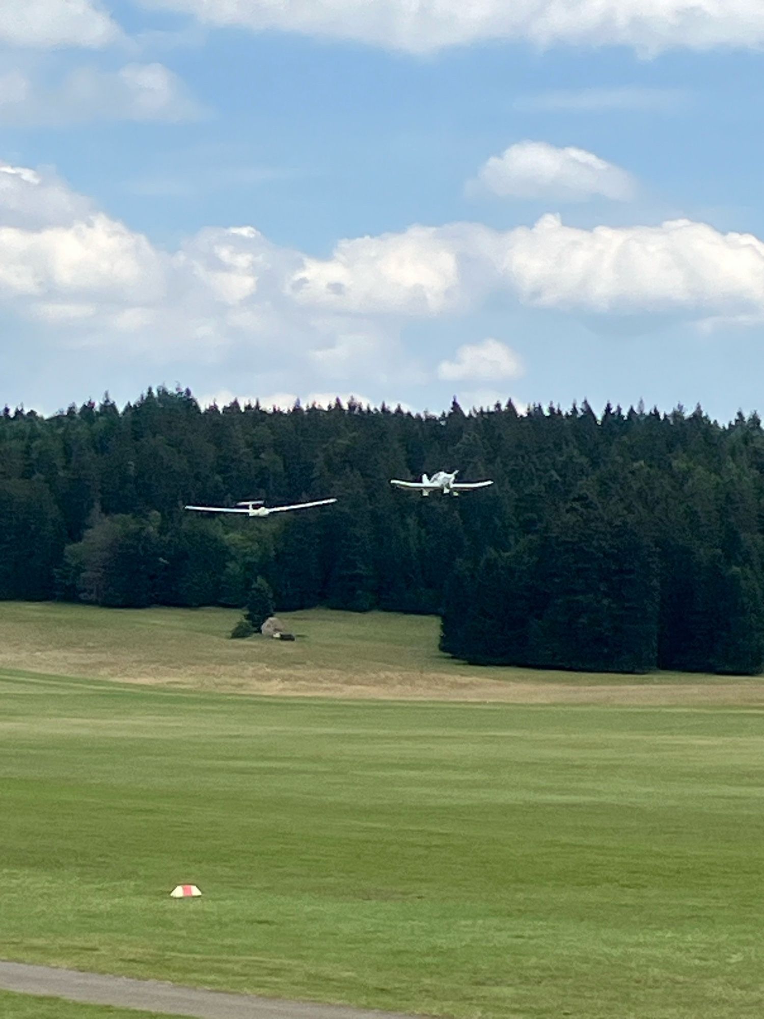 Hockantfoto: ein Segelflugzeug, das von einem Motorflugzeug gezogen wird. Im Hintergrund sieht man einen Wald und am Waldrand steht eine Kapelle. Unten ist grüne Wiese und ein rot-weißes Hütchen, das die Landebahn markiert. Oben ist der Himmel teilweise bewölkt.