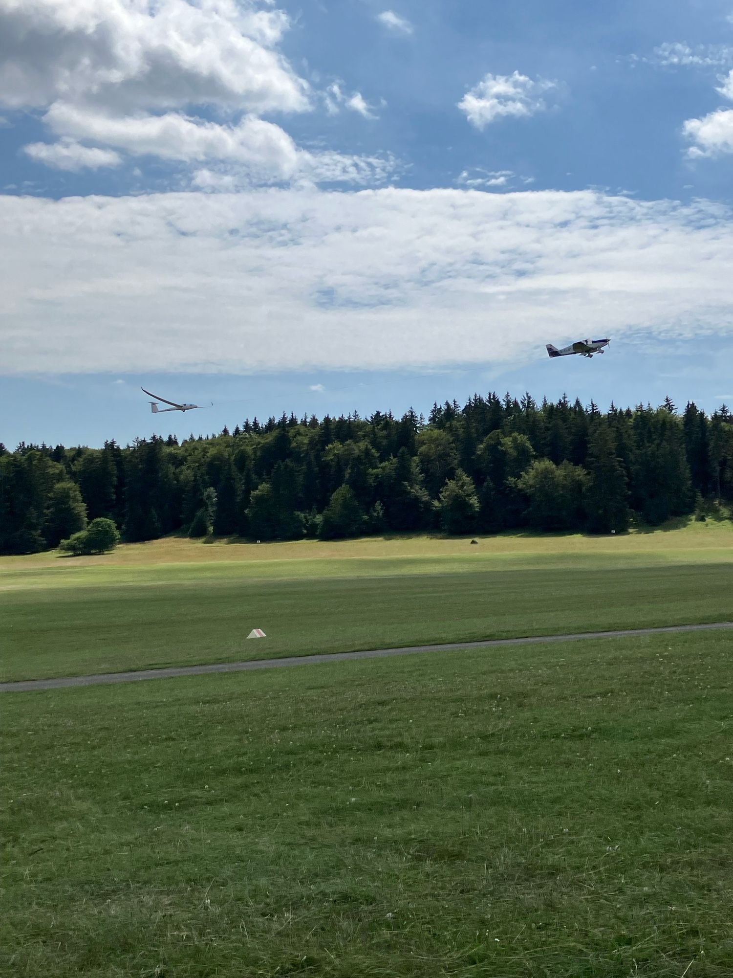 Hochkantfoto: ein Segelflugzeug wird von einem Motorflugzeug gezogen (geschleppt), sie fliegen bereits über den Bäumen, die man im Hintergrund sehen kann. Unten ist grüne Wiese, oben leicht bewölkter Himmel