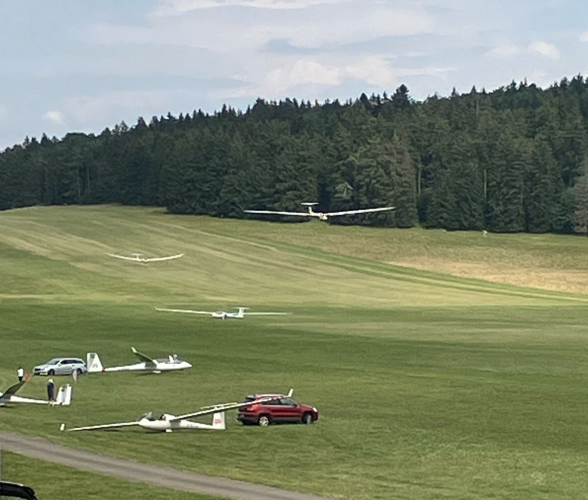 Vorne links im Bild werden zwei Segelflugzeuge von je einem Auto von der Wiese gezogen. Direkt dahinter rollt ein gerade gelandetes Segelflugzeug nach links weg. Weiter hinten sind zwei weitere Flugzeuge im Landeanflug. Ganz hinten steht ein dunkler Wald, oben drüber ist der Himmel recht voll mit Wolken