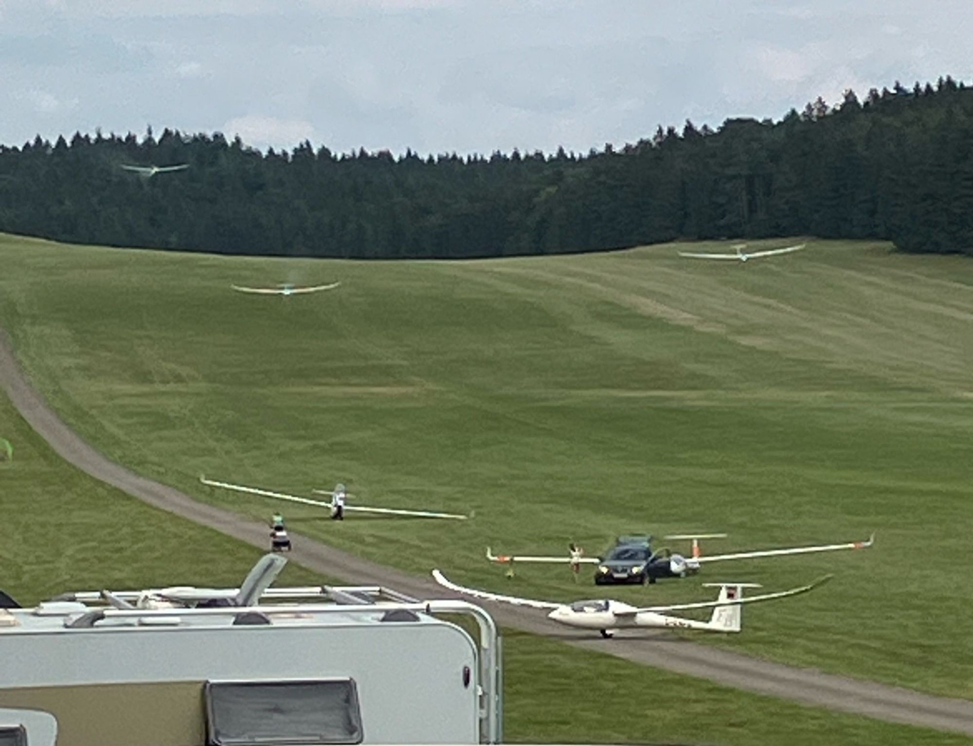 Im Vordergrund links sieht man den "Popo" von einem Wohnmobil, direkt dahinter stehen drei Segelflugzeuge am Boden. Eines davon wird von einem Auto gezogen im Hintergrund sieht man auf unterschiedlicher Höhe drei weitere Segelflugzeuge im Landeanflug. Weiter hinten ist ein dunkler Wald, darüber ein leicht grauer Himmel