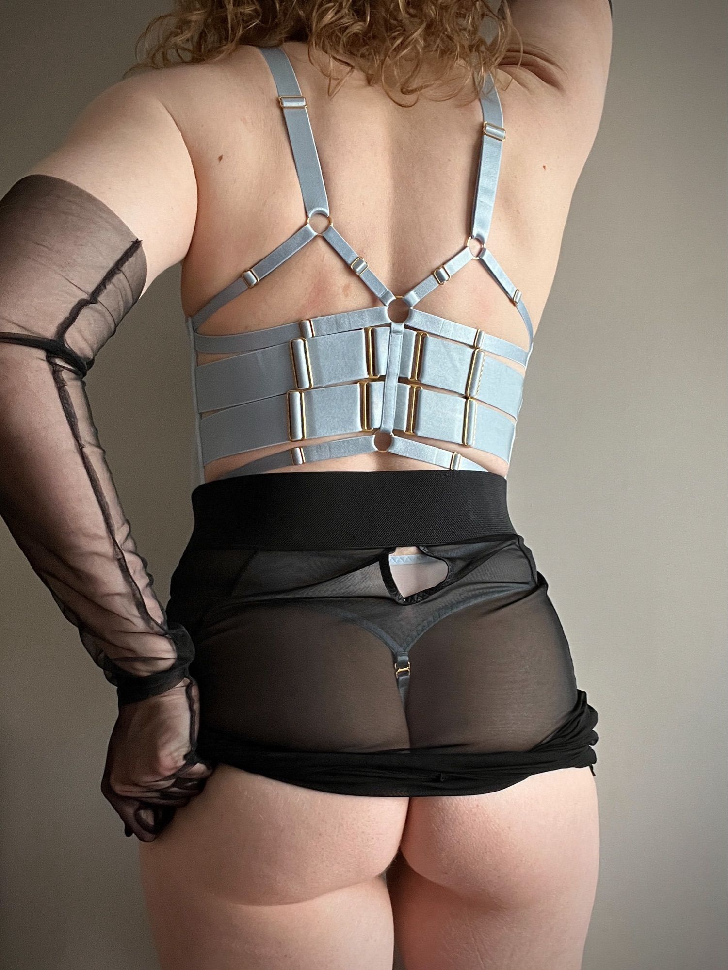 Photo of me from behind wearing a Bordelle dusty blue strappy bodysuit, Edge of Beyond black mesh pencil skirt and black mesh opera length gloves. I’ve hitched the skirt up with one hand to reveal half my ass…who’s getting on their knees?