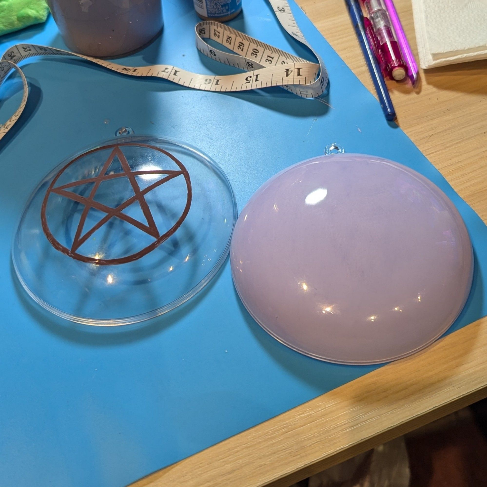 Crafting workspace featuring two halves of an acrylic ornament. One is paint a solid lavender color and the second has a brown pentagram painted on top.