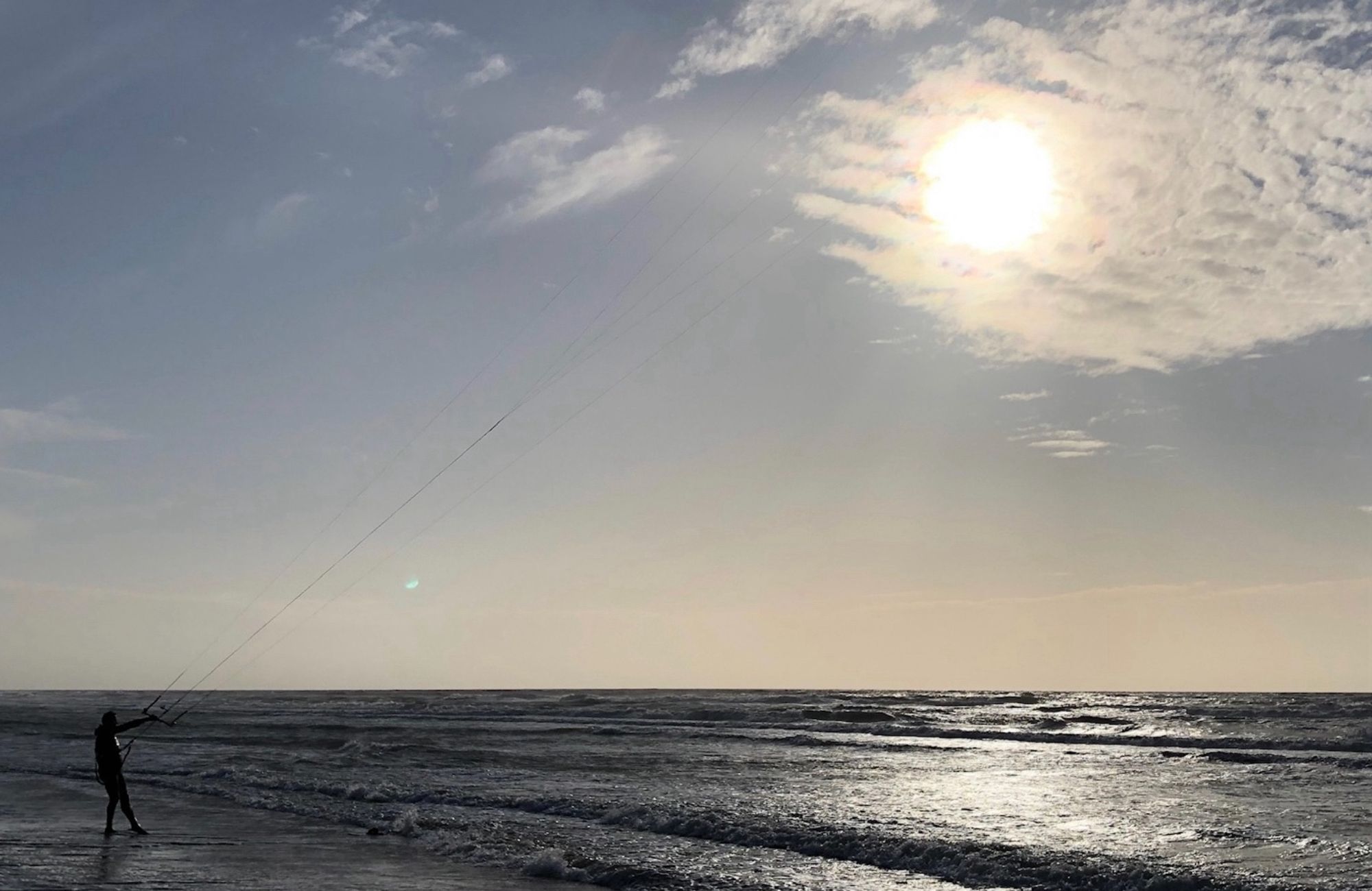 Strand, zee, koude zon, op de voorgrond een vliegeraar met de lijnen in zijn hand, je ziet de vlieger niet, de lijnen leiden als het ware naar de zon, zo lijkt het dat hij vliegert met de zon