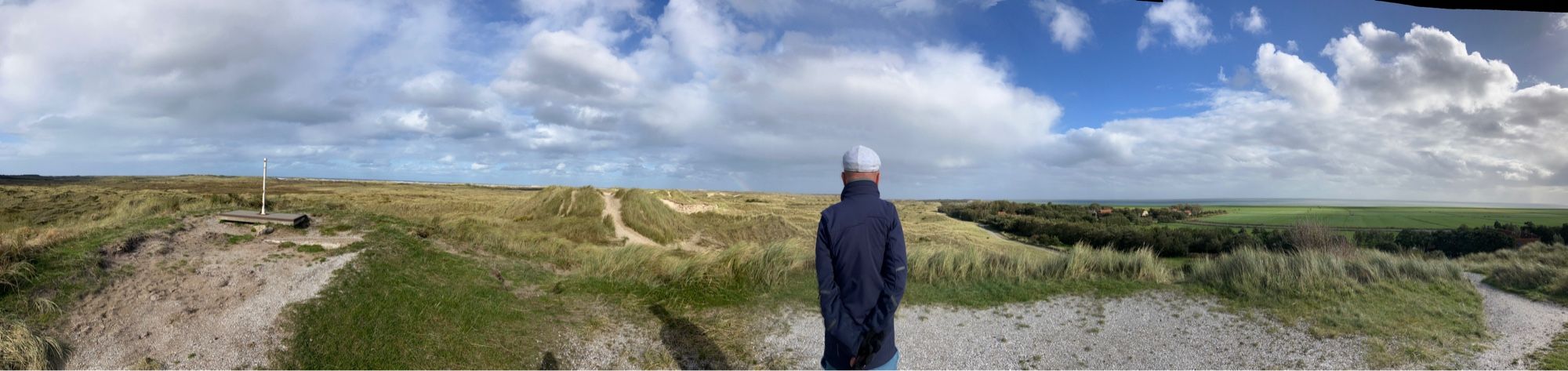 Panorama beeld met helemaal links de Noordzee en rechts Oosterend met de wadden, geel groene toppen van de duinen en in midden man mat handen in de zakken en lichtblauw petje op