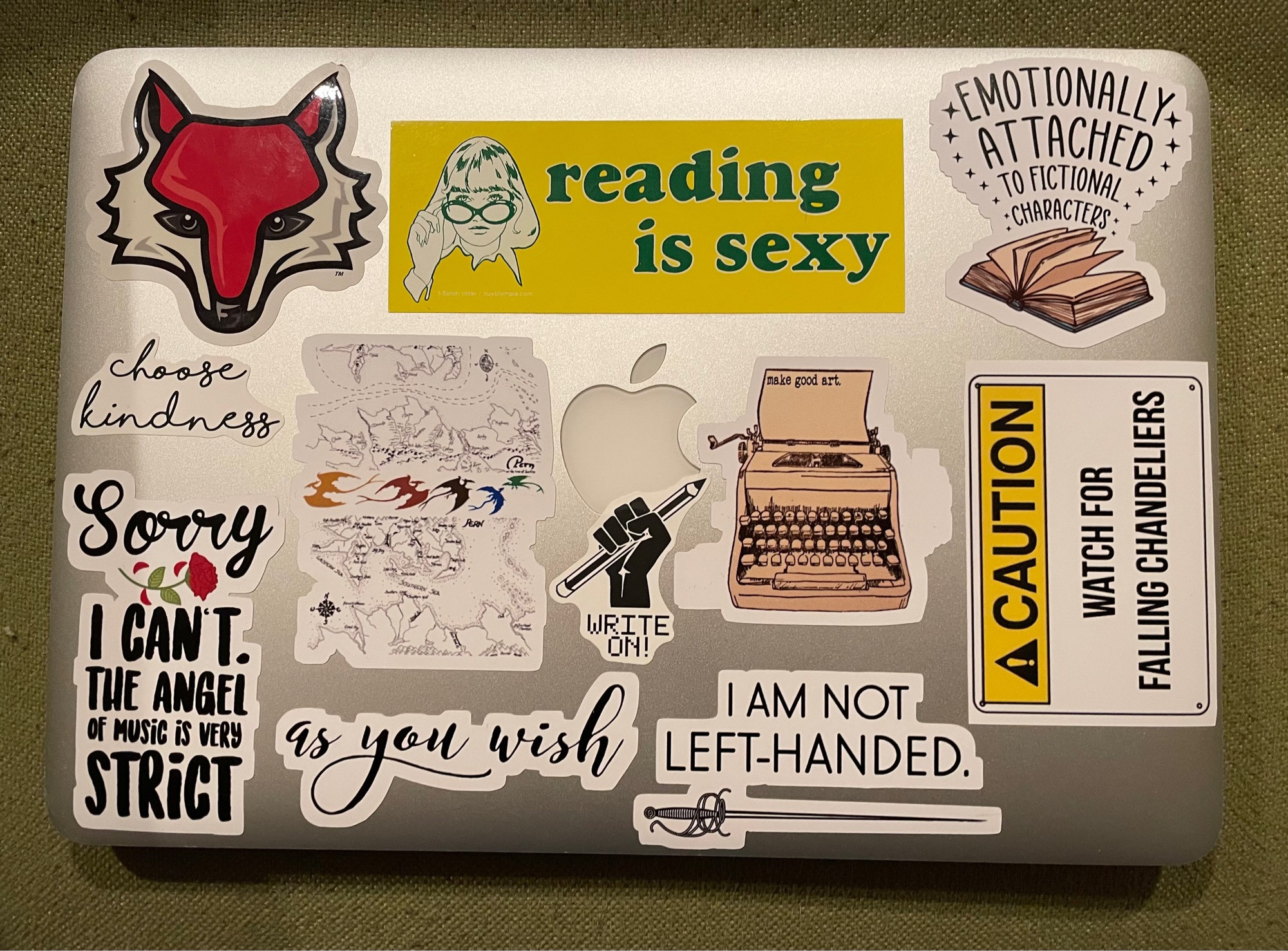 An Apple laptop covered in writing-related stickers: Reading is sexy, emotionally attached to fictional characters, as you wish, I am not left handed, write on, make good art, sorry I can’t the angel of music is very strict, caution watch for falling chandeliers, choose kindness