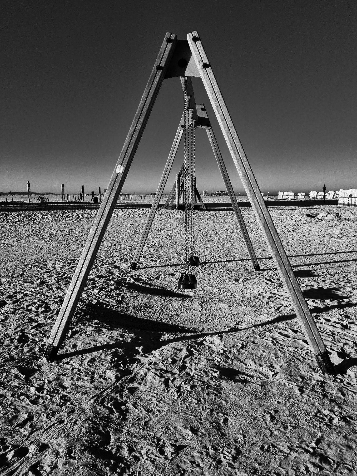 Ein Schaukelgerüst aus Holz mit zwei Schaukeln am Strand. Im Hintergrund ist ein Holzsteg zu sehen, rechts im Hintergrund sind auch Strandkörbe zu sehen.