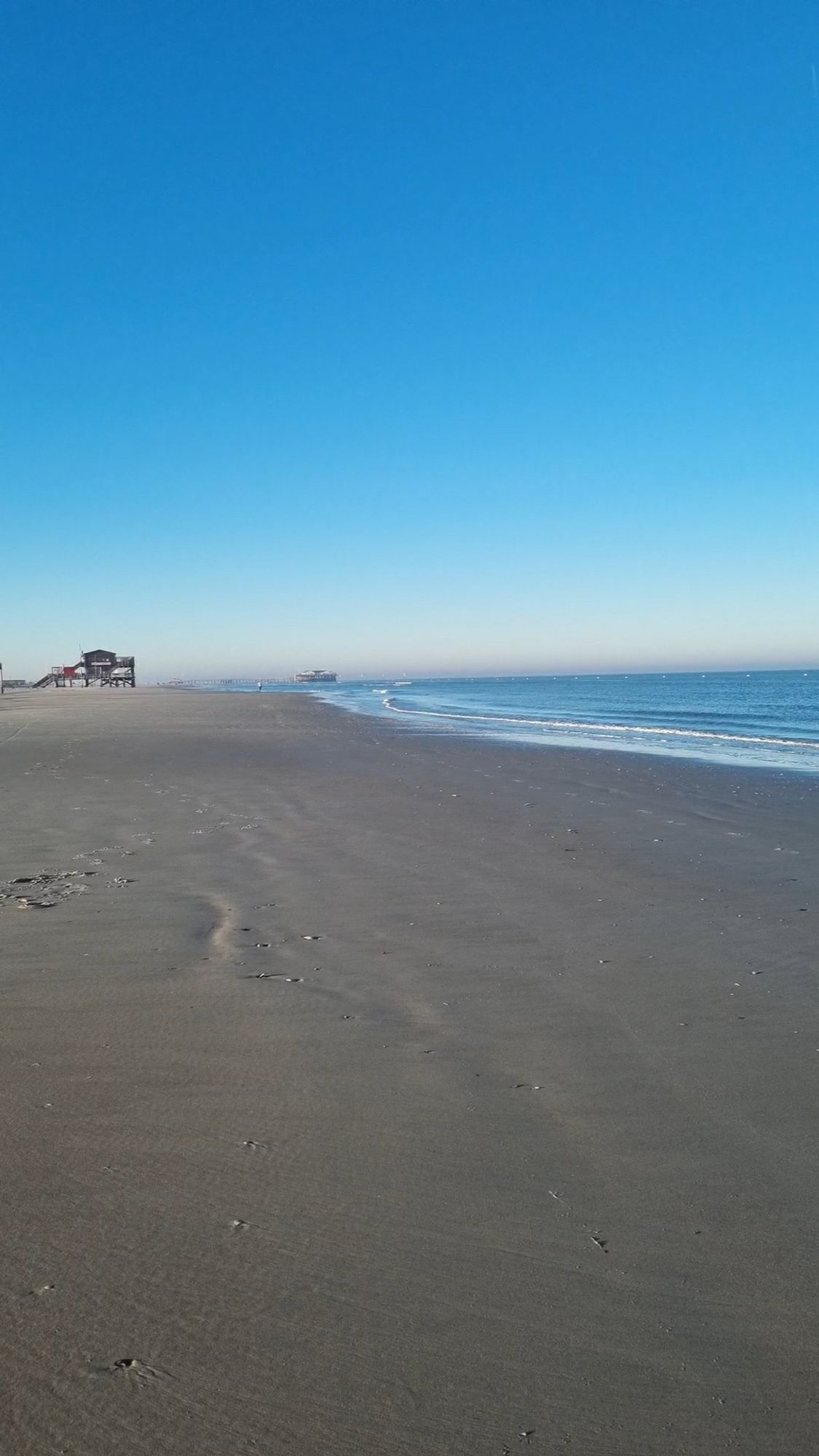 Den unteren Teil des Bildes mit Sandstrand ein ein, eine kleine Ecke rechts das Meer. Am Horizont ist links in der Ferne ein Pfahlbau zu sehen. Der obere Teil des Bildes ist hellblauer, wolkenloser Himmel.