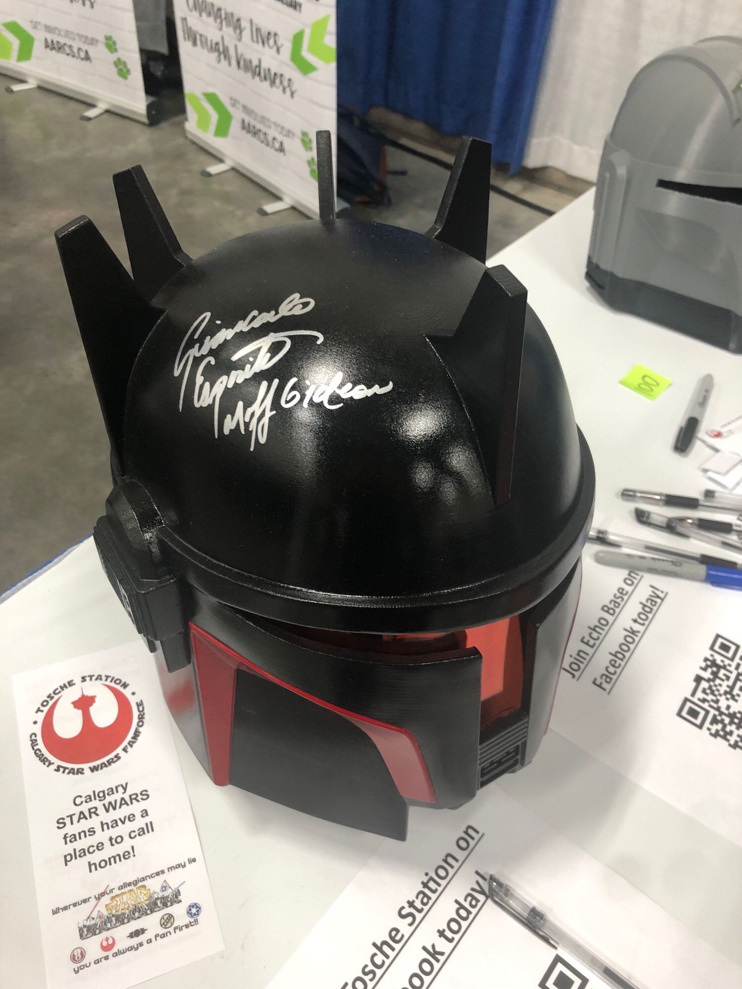 A black helm with red highlights and a T-shaped visor in the Mandalorian style. It’s autographed in silver sharpie, the inscription reading: “Ginacarlo Esposito, Moff Gideon.”