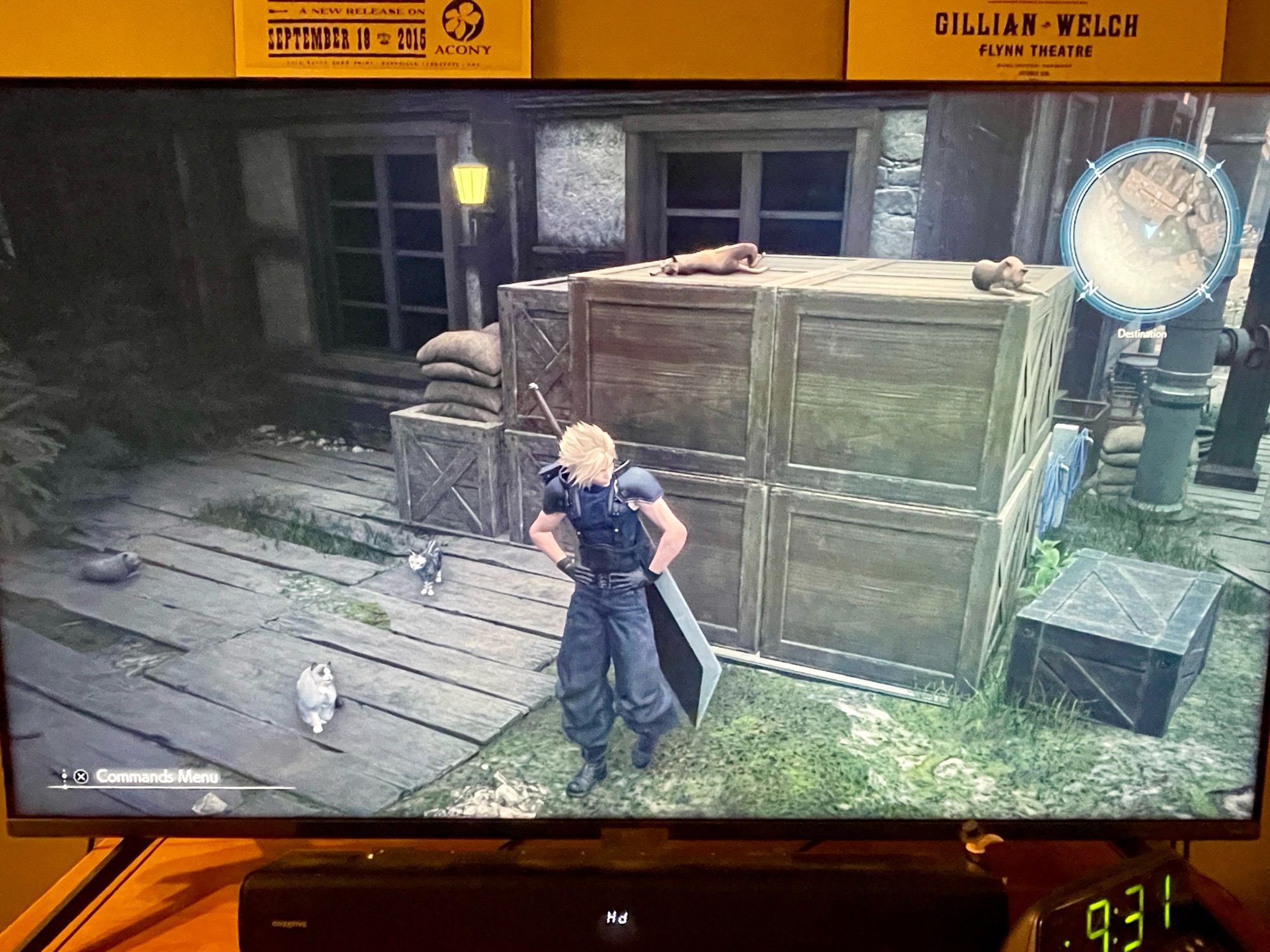 A photo of my tv screen. The character Cloud Strife stands in the center of a back alley with crates and such piled up behind him. There are three cats lounging on the ground to his right and on top of the boxes are two more, one of which is all stretched out on its back. The cat animation is pretty dang good.