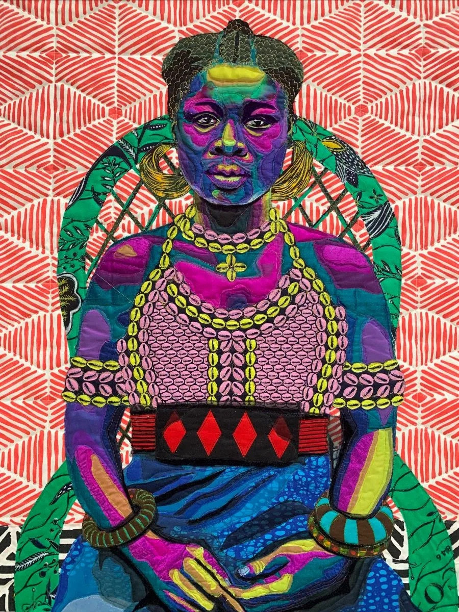 A quilt portrait of a striking young woman. The background fabric is red and cream geometric stripes. The girl is seated in a green chair and is sitting with her hands lightly resting in her lap. She states confidently ahead, straight at the viewer. She wears a bodice of cowrie shells and has beautiful bangles on each wrist. Her Black skin is depicted in a gorgeous array of pinks, purples, blues, and lime green