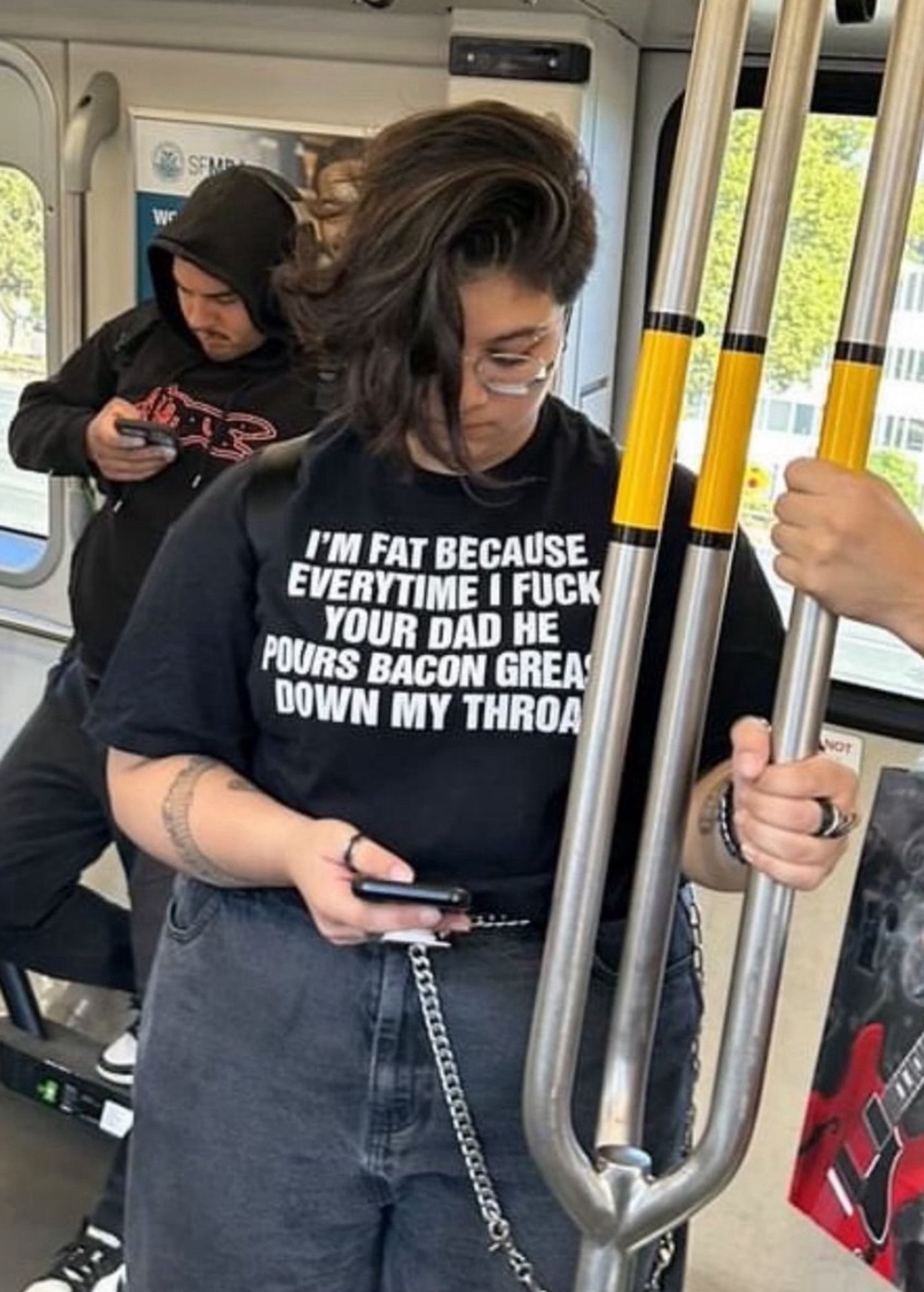 A person standing in a subway car wearing a black t-shirt that reads: "I'm fat because every time I fuck your dad he pours bacon grease down my throat"