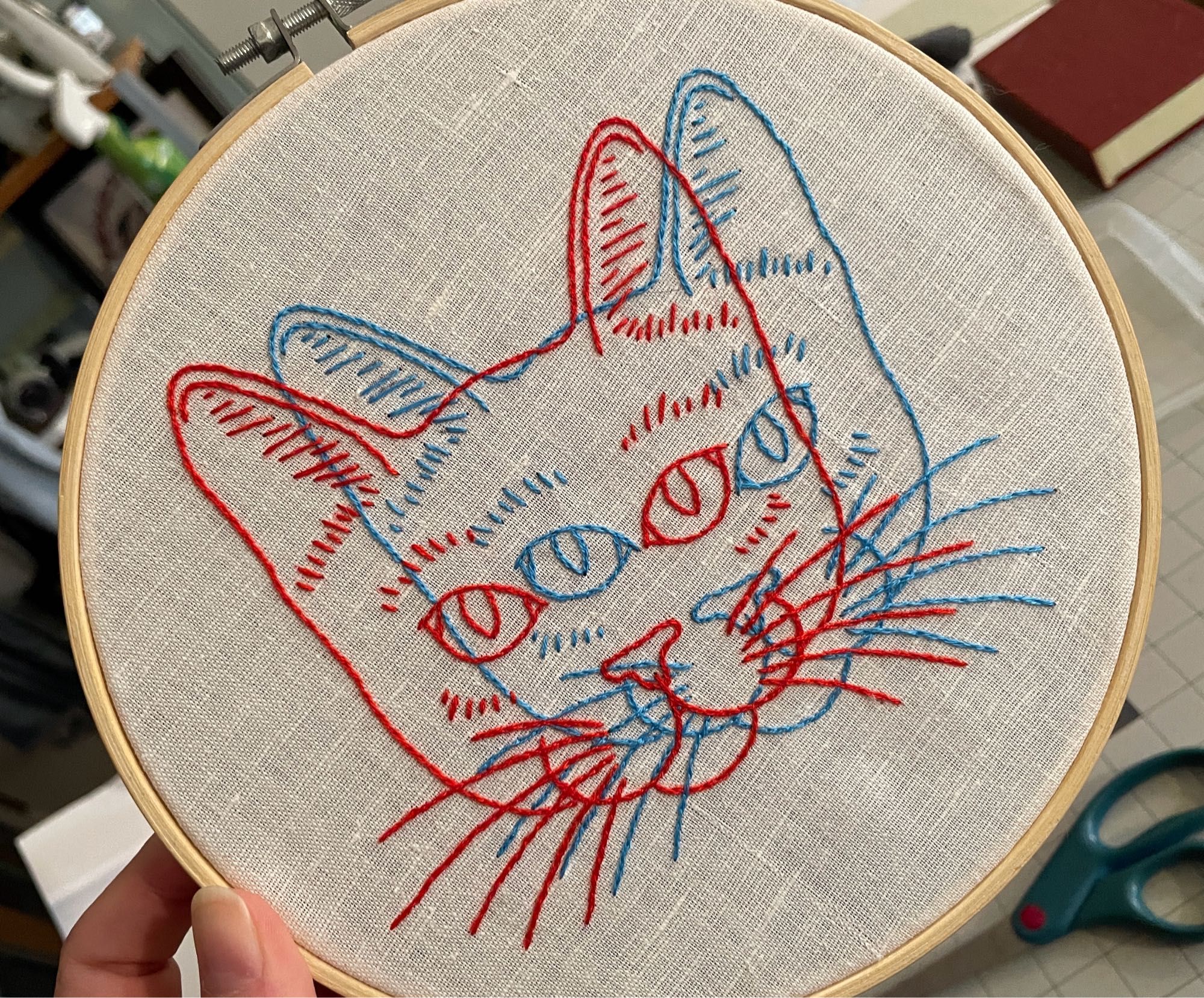 An embroidery in a hoop on white linen. It’s a double image of a cat face, slightly offset from one another. One is done in red and one in blue like an old school 3D glasses movie