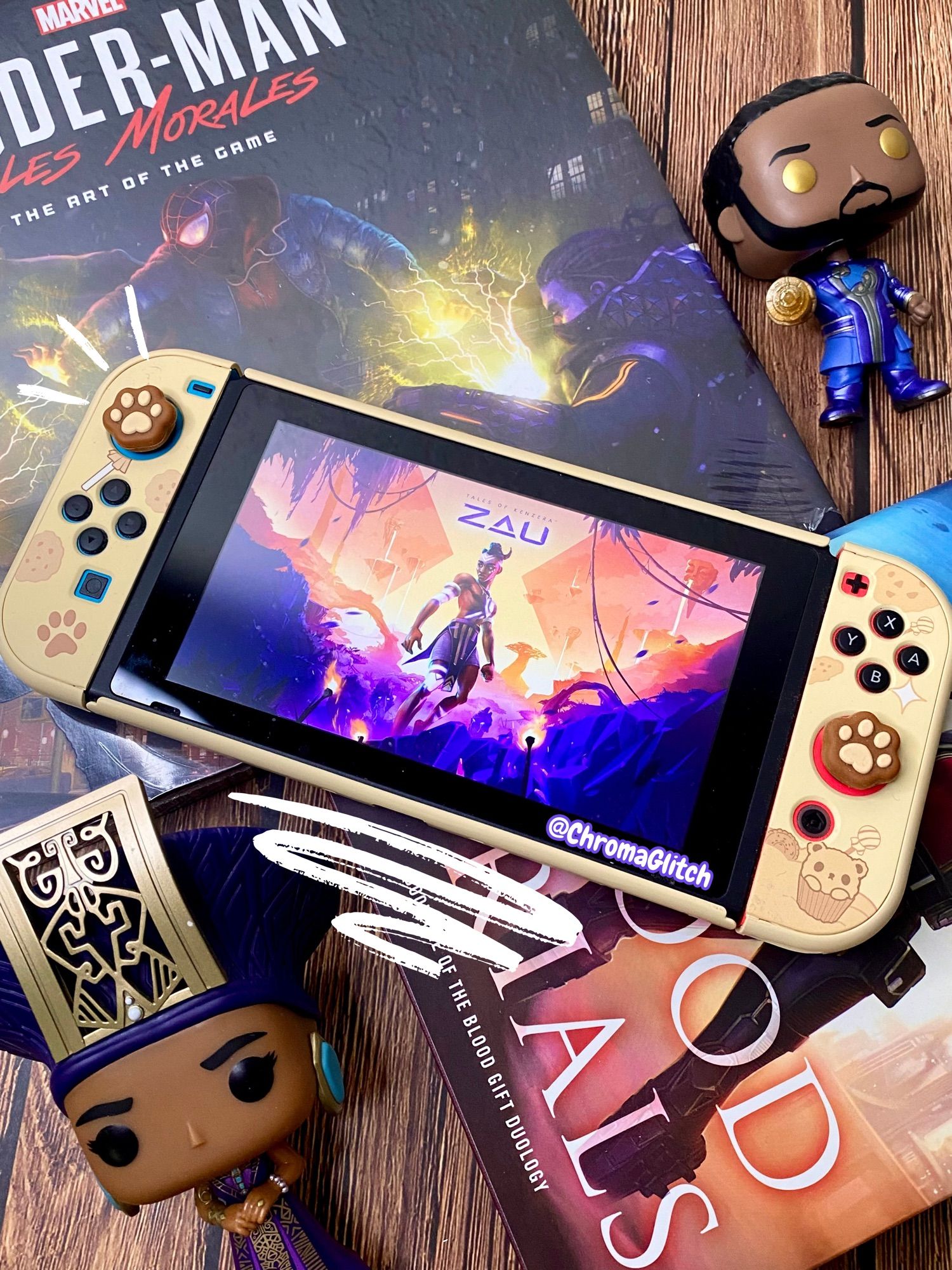 Image of a flat lay with a Nintendo Switch in the center, surrounded by books and funko pops. On the screen is the cover art for The Tale of Kenzera: Zau.