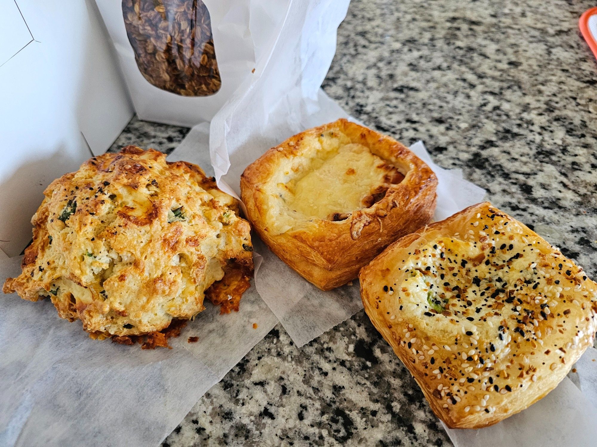 A collection of savory baked goods including: a cheddar scallion scone, a croque monsieur, an everything bagel danish, and a bag of house made maple pecan granola in the back