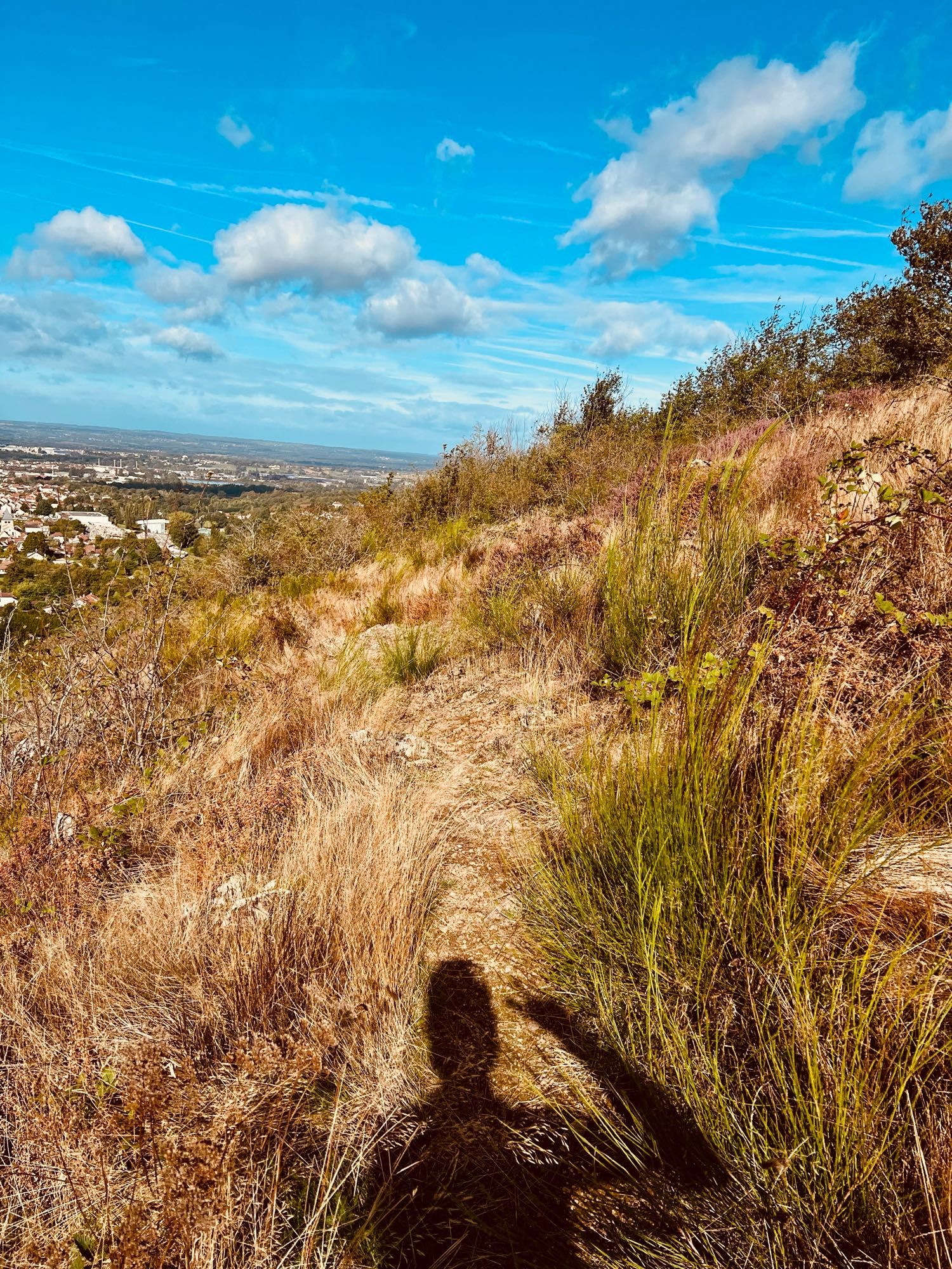 Un sentier rocailleux très étroit, à flanc de corniche sur un sol de lande, avec des herbes basses. Au fond, le ciel est bleu, avec quelques nuages de beau temps. En contrebas sur la gauche, la ville, que je ne v us pas rejoindre tout de suite.
Au premier plan, l’ombre de ma tête et de ma main droite qui fait un V. Pas pour la perf du jour mais parce que j’adore ce passage.