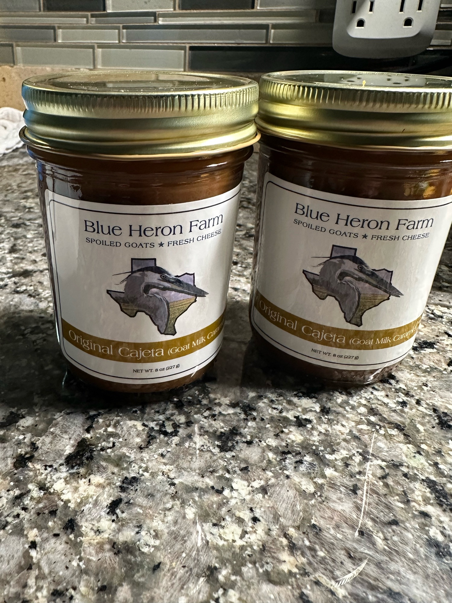 Two jars of Blue Heron Cajeta (goat milk caramel) sit on a grey granite countertop. The label beautiful with a blue heron inside the state of Texas. 