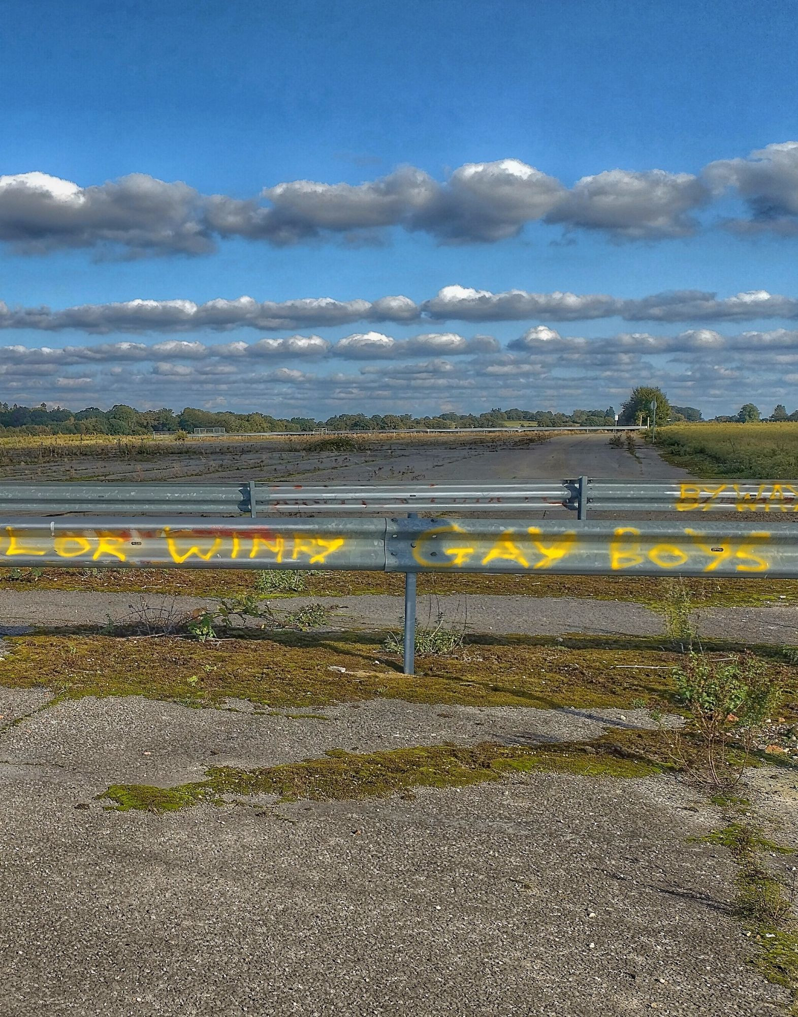 Metal barrier across airfield runway, left half of pictured graffitied in yellow with "Taylor Wimpy Gay Boys" 