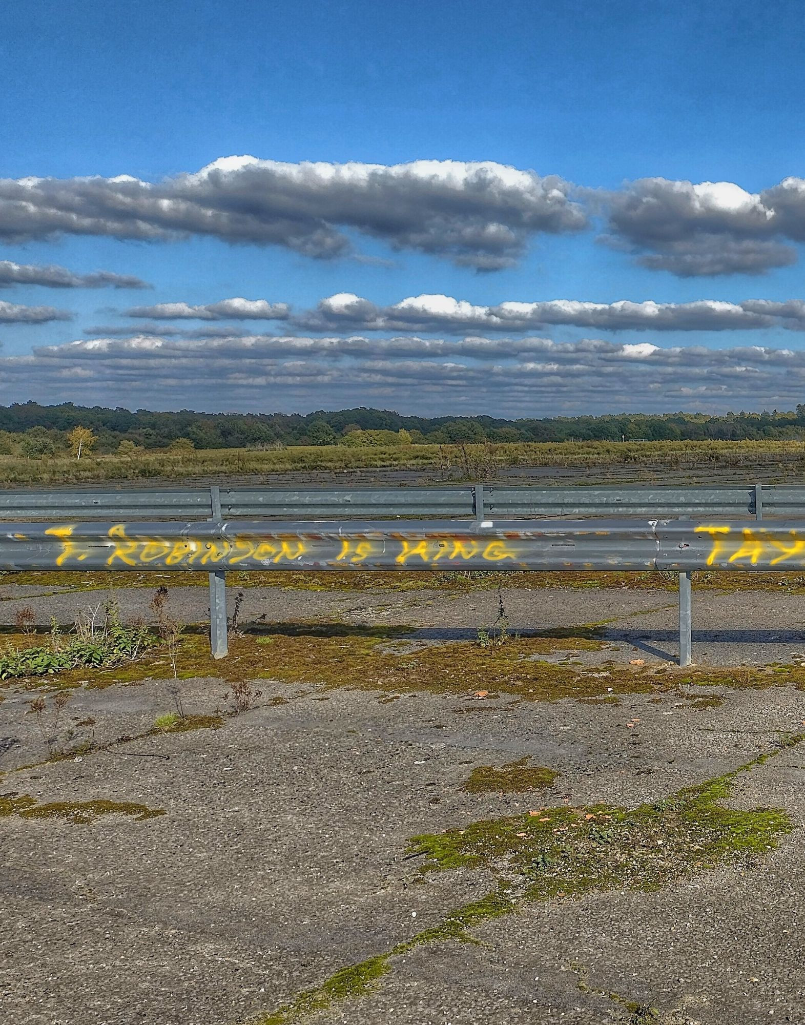 Metal barrier across airfield runway, left half of pictured graffitied in yellow with "T. Robinson is king" 