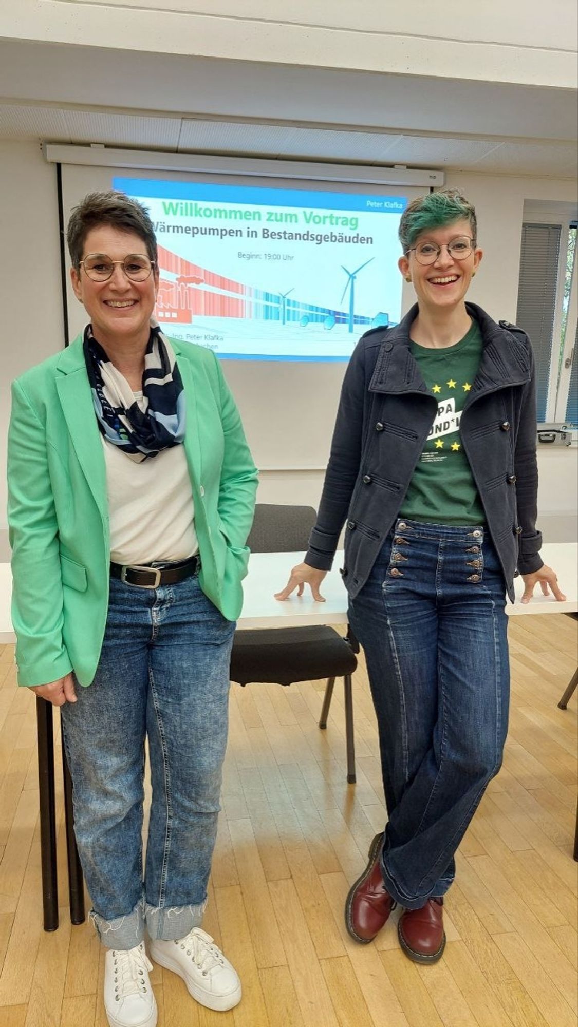 Eine kurzhaarige Frau mit Brille, Jeans und grünem Blazer neben mir (Brille, Jeans, grünes Shirt und blaue Jacke) vor einer Vortragsfolie zu Wärmepumpen in Bestandsgebäuden