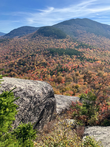 View from rocky outcrop onto another mountain filled with colorful forests and topped with dark green conifers