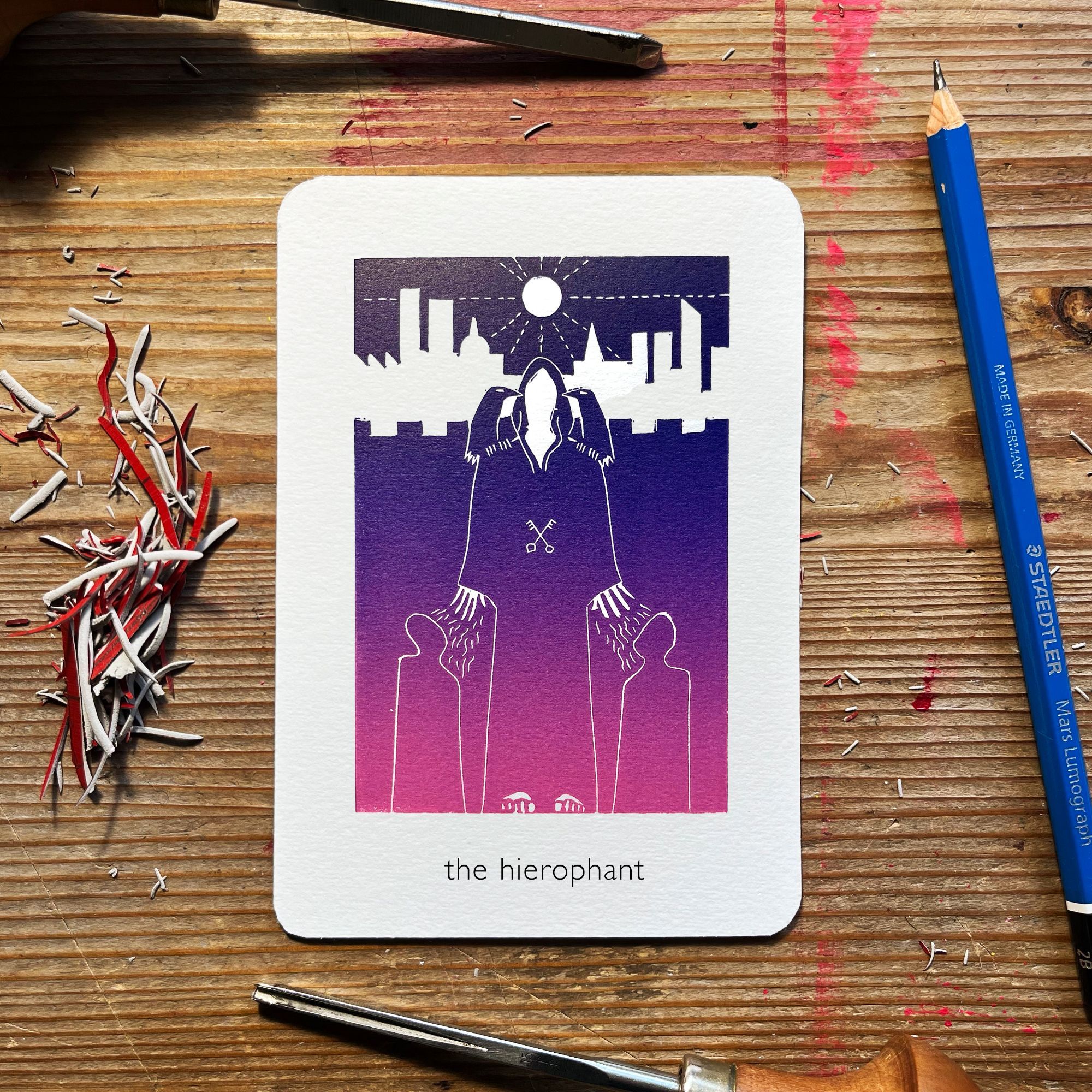 a linoprinted tarot card surrounded by linoprinting tools and offcuts. It's printed with a gradient of deep purple to rich pink. at the top is a sun or full moon emitting rays. below that is a cityscape horizon line. Before that is a castellated wall. In the foreground is a monk stood with their hood up. on each shoulder is a crow. there are crossed keys at their solar plexus. kneeling in front of them are two figures. waves of energy come from the monk's spread fingers towards the kneeling figures' faces.