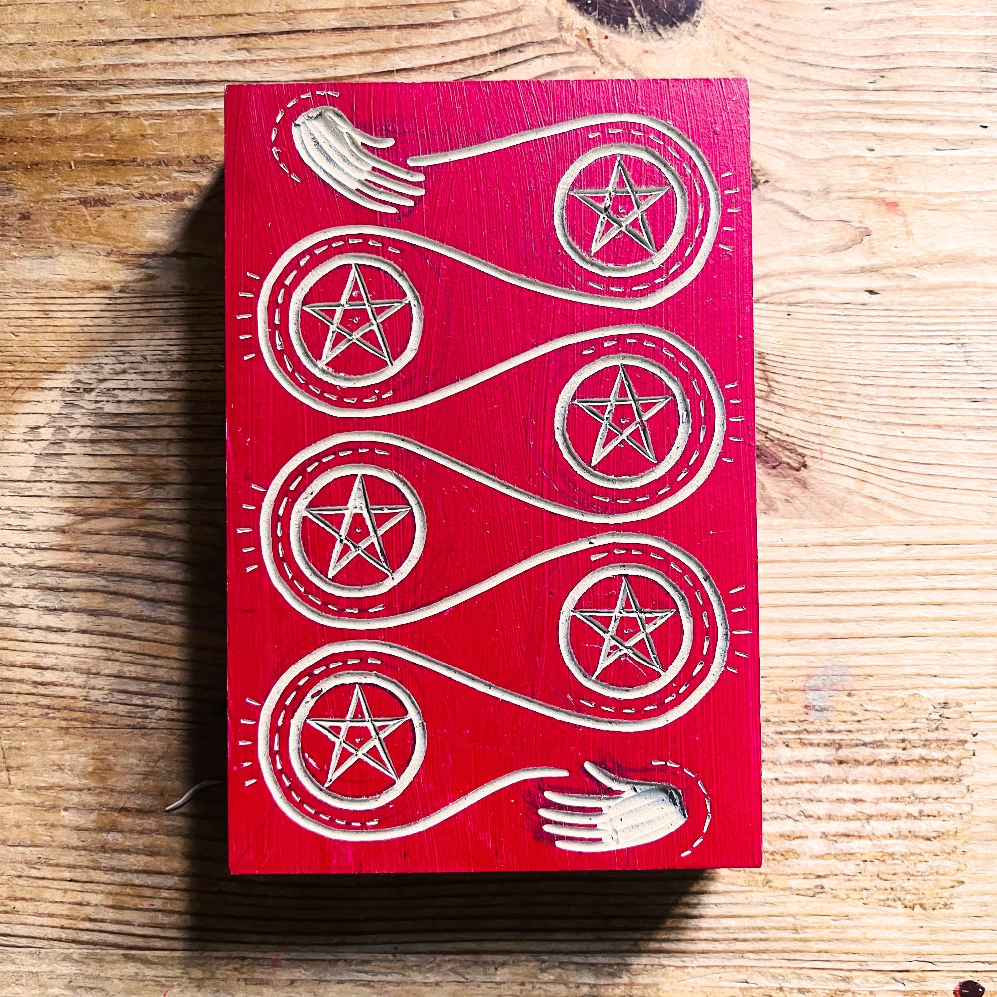 a cut lino plate that's been painted red. There's a hand at the top and a hand at the bottom. From top to bottom is a looping meandering line and at each loop is a pentacle.