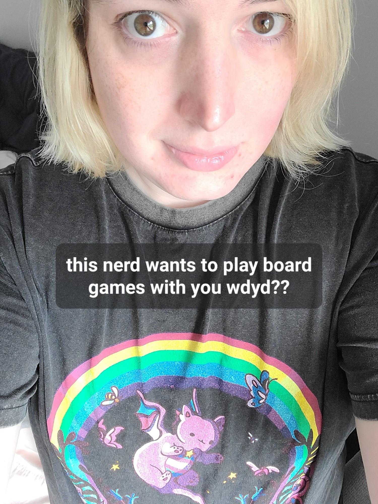 selfie of an enby with blonde bleached hair looking slightly up at the camera and wearing a shirt with a rainbow and trans colors. a caption reads "this nerd wants to play board games with you wdyd??"