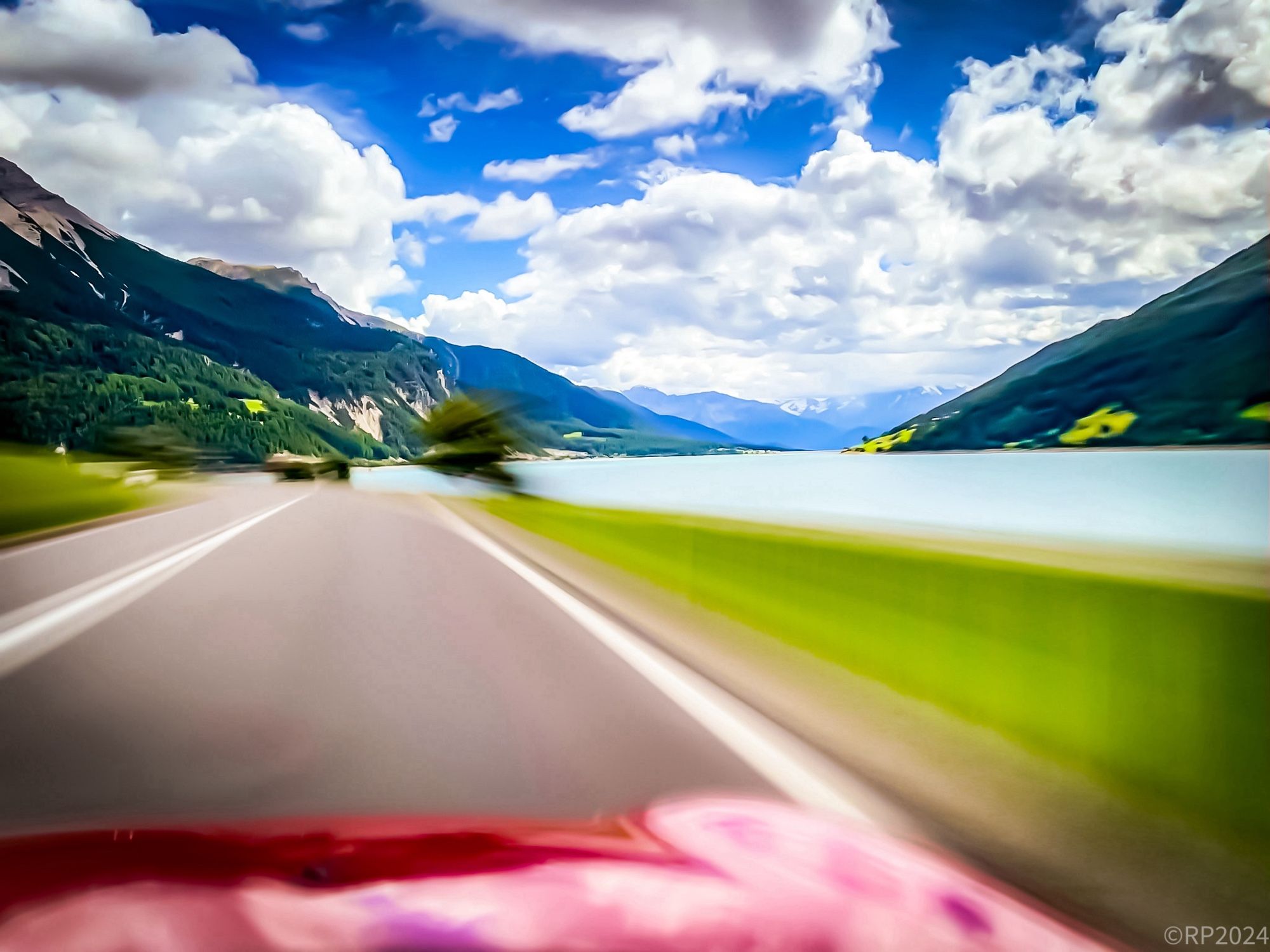 Strasse durch das Vinschgau. Strasse, Auto und Fahrbahnrand sind dynamisch verwischt. Berge und Himmel sind klar.