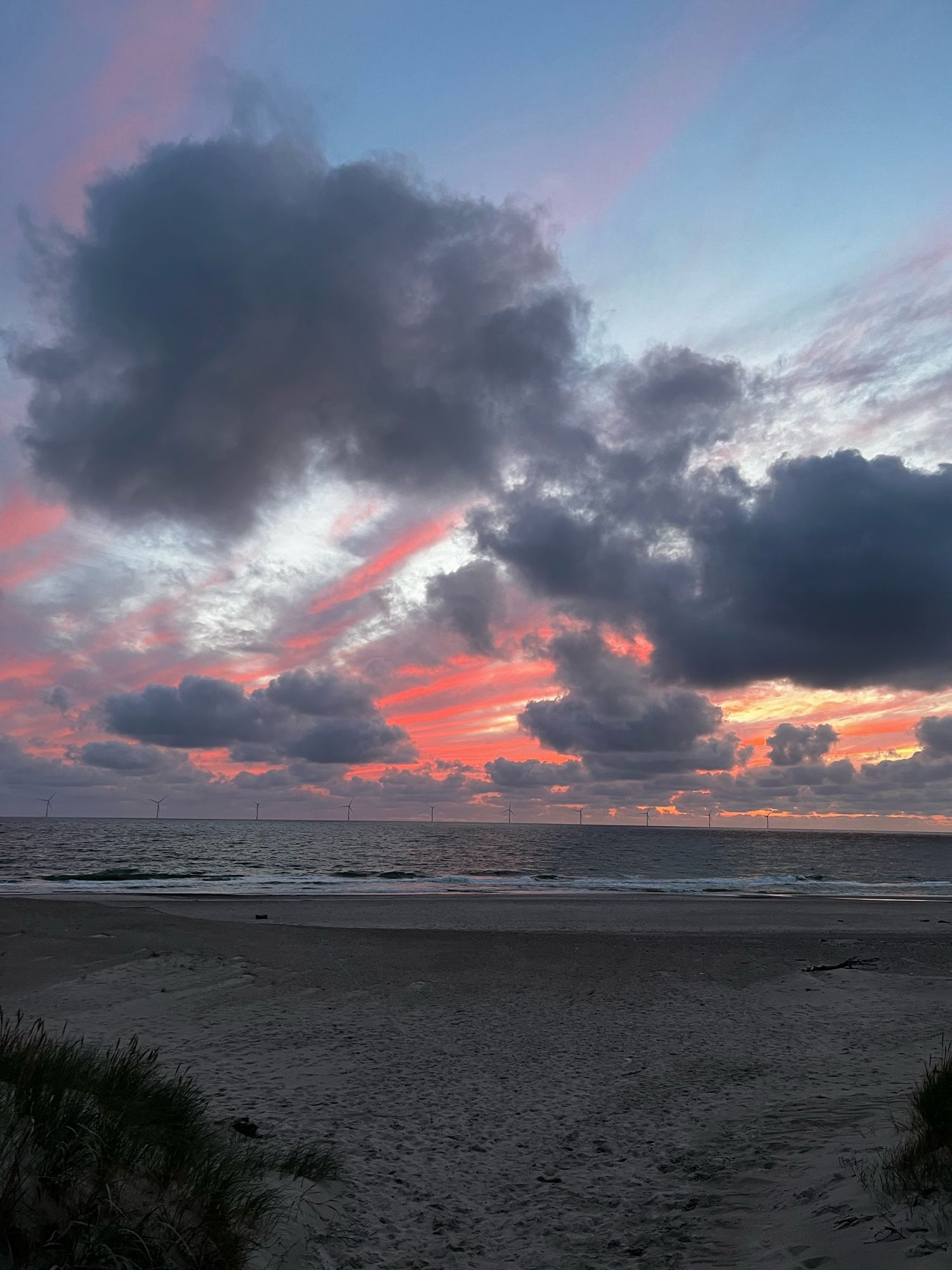 Sonnenuntergangsstimmung am Meer. Unten ein wenig Dünengras. Strand und Meer mit kleinen Wellen. Der Himmel ist wolkig, orange, grau und gelb.