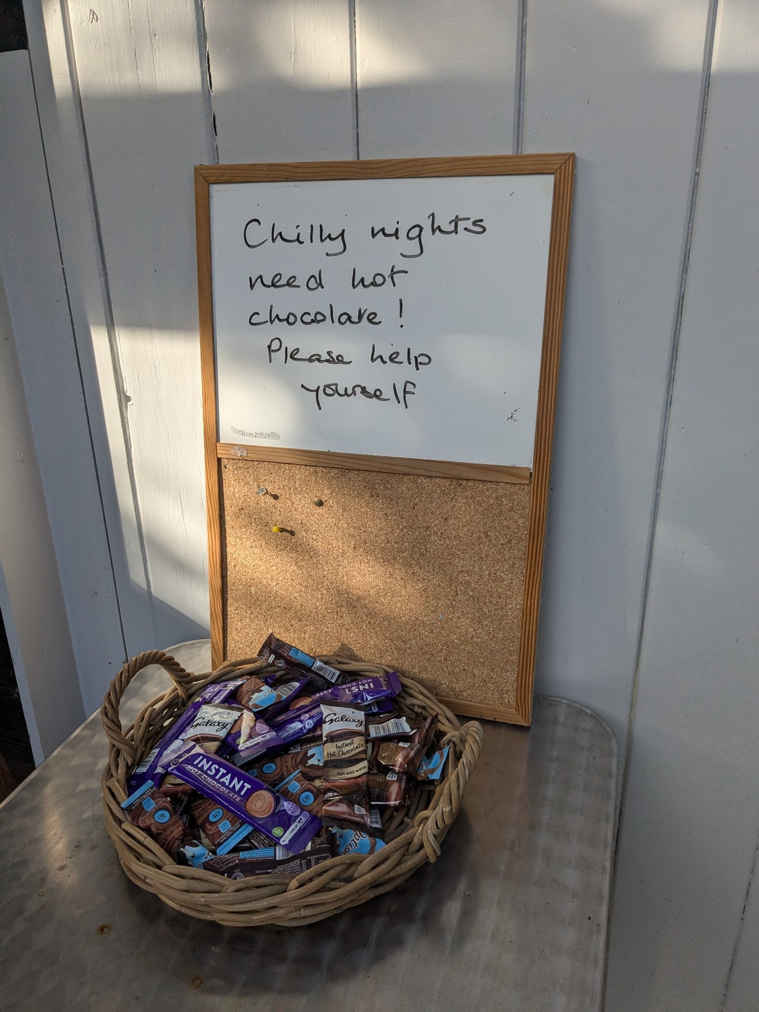 A basket of hot chocolate sachets in front of a noticeboard that says "Chilly nights need hot chocolate! Please help yourself"