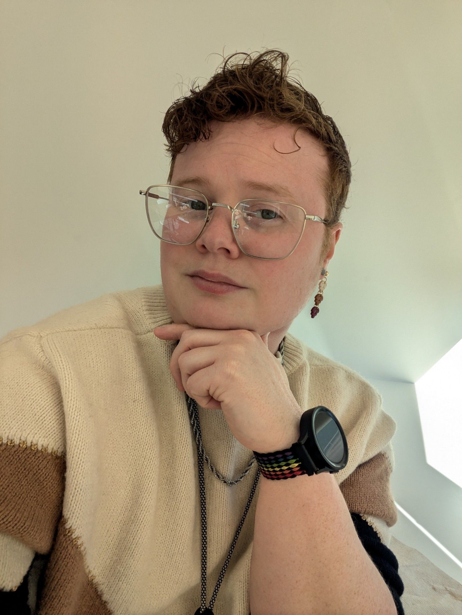 A selfie of nonbinary person wearing autumn colored sweater, white orange and red leaf wearings, a pride smart watch, and large rectangle glasses.