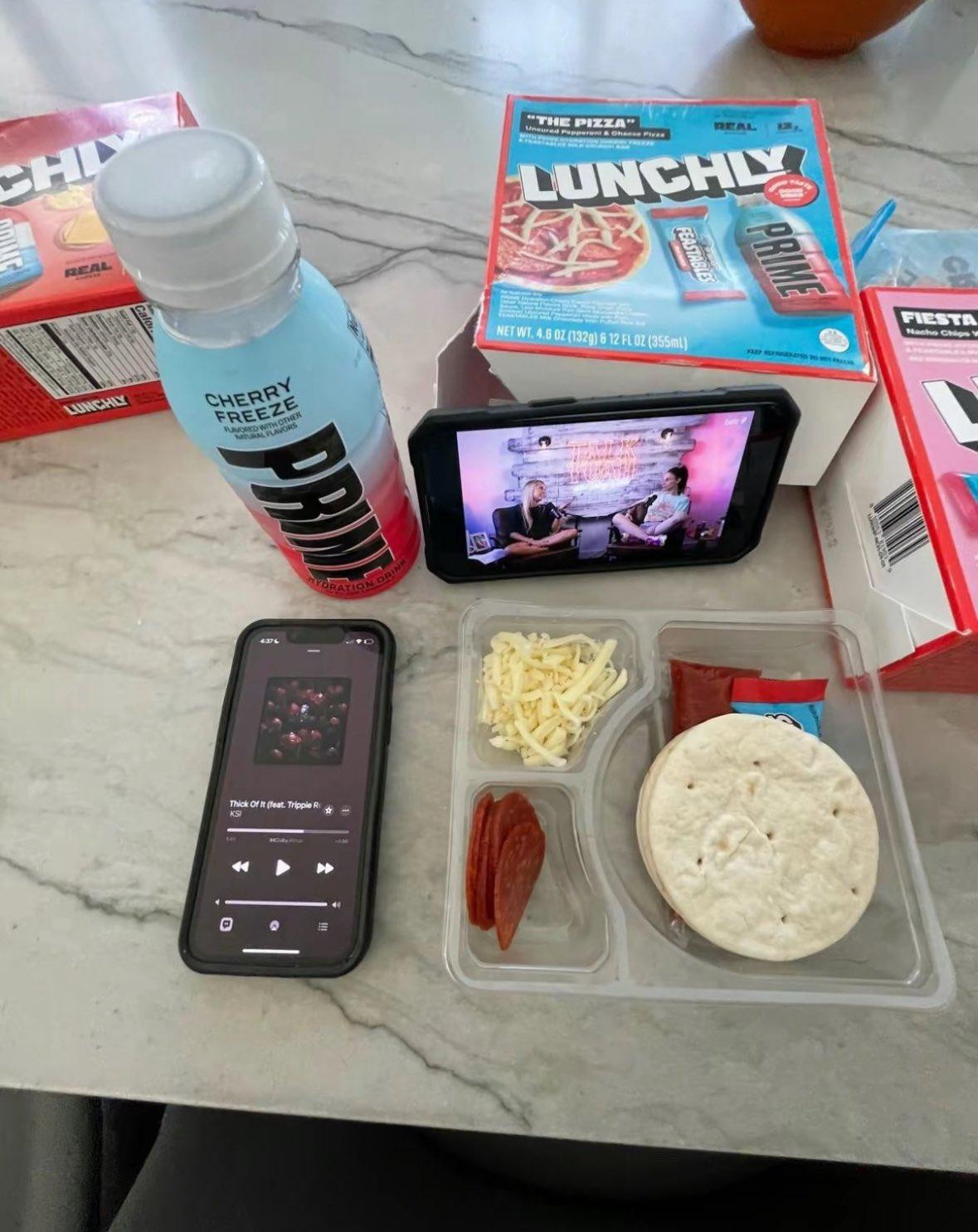 a marble table with a few boxes of Lunchly sitting around, a phone is propped up against one of the boxes showing the Talk Tuah with Haliey Welch podcast and there's a packet of Lunchly "the pizza" flavour, a bottle of cherry freeze prime, and a phone playing Thick of It by KSI