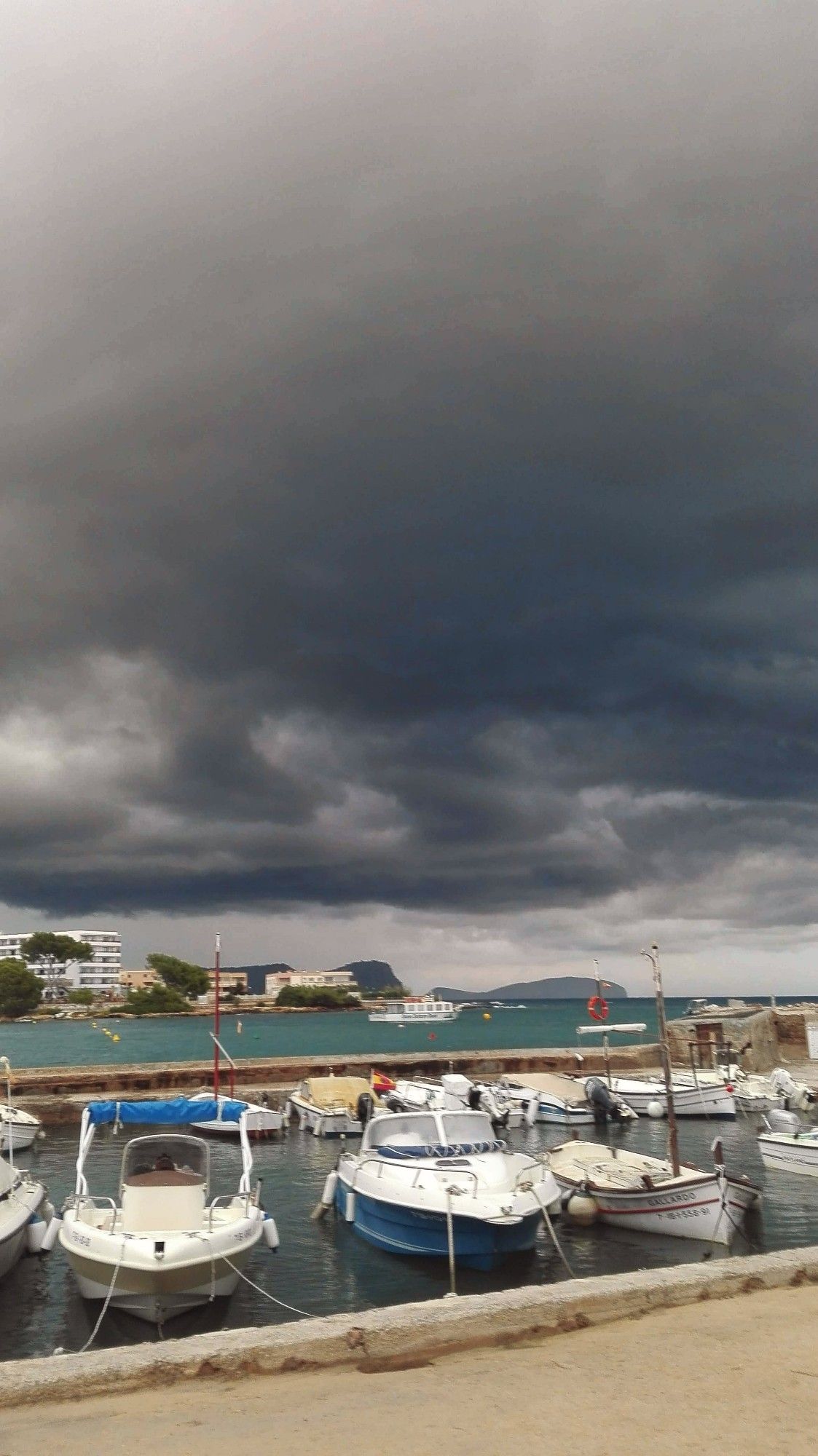 Ein Hafen mit kleinen Booten. Im Hintergrund ein Hotel. Über allem der Himmel mit bedrohlich aussehenden dunklen Wolken.